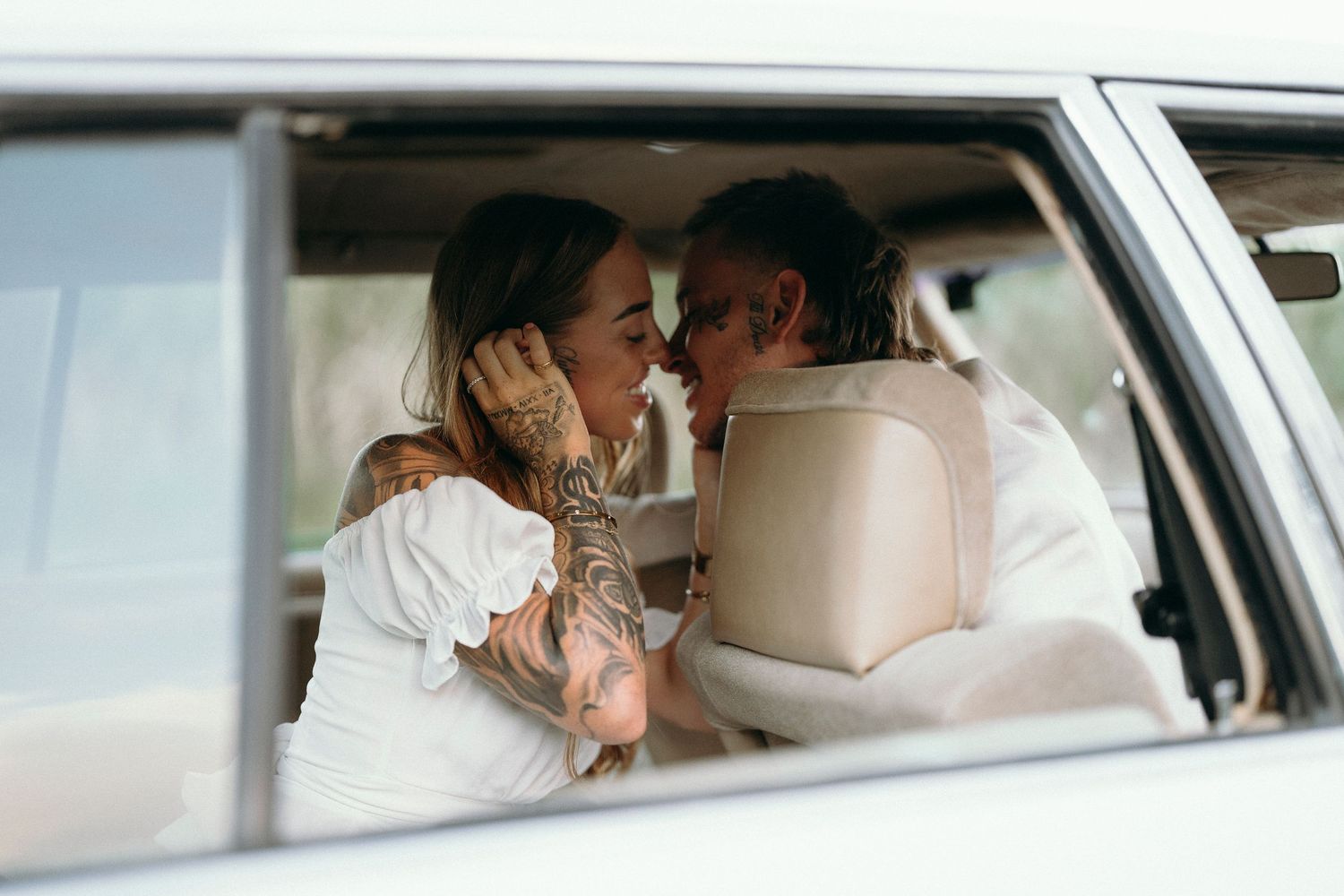 Tattooed couple sharing a romantic moment inside a vintage white car, leaning in close for a kiss.