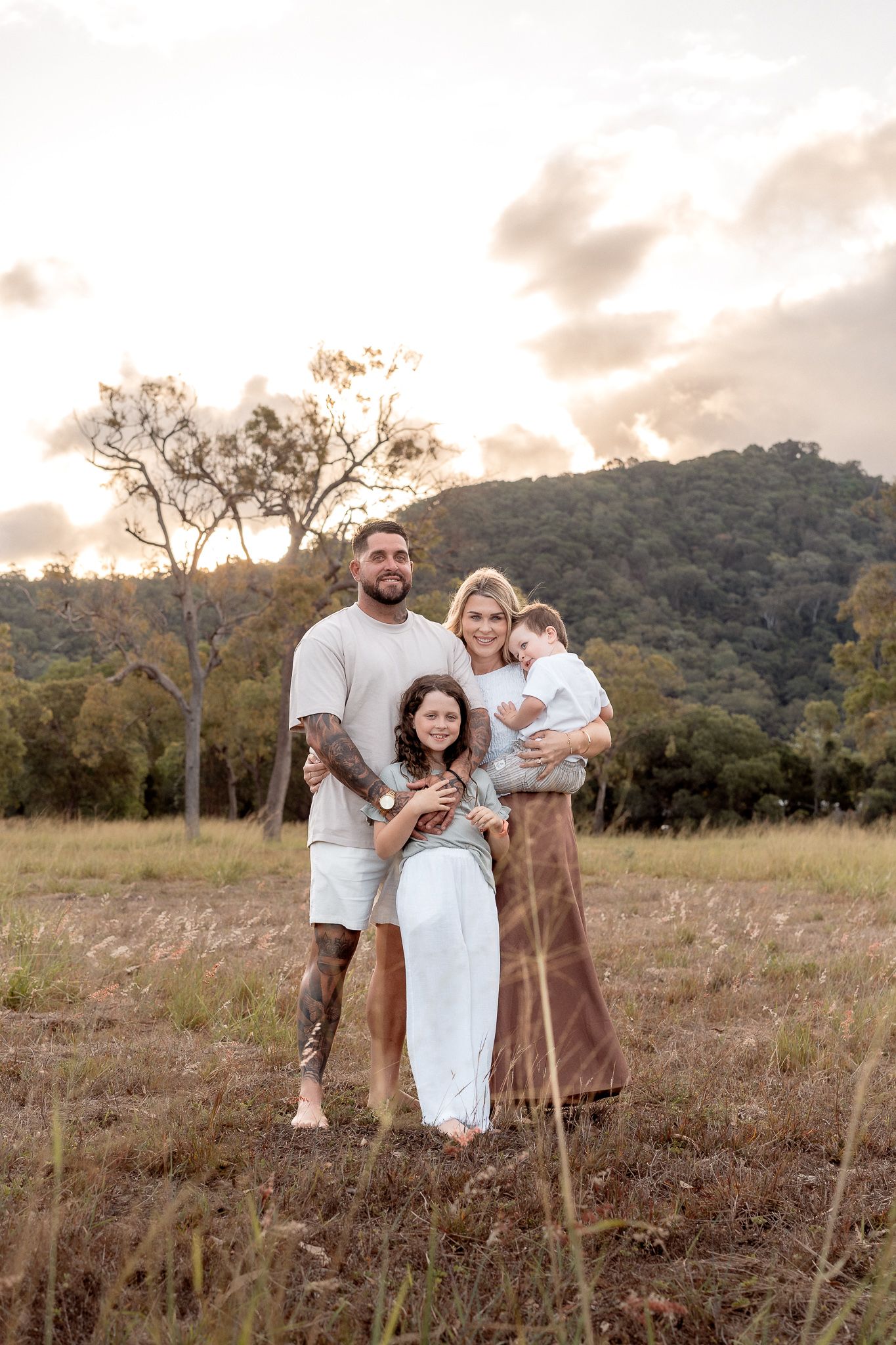 Happy family of four posing together in golden field at sunset with forested hills in background.