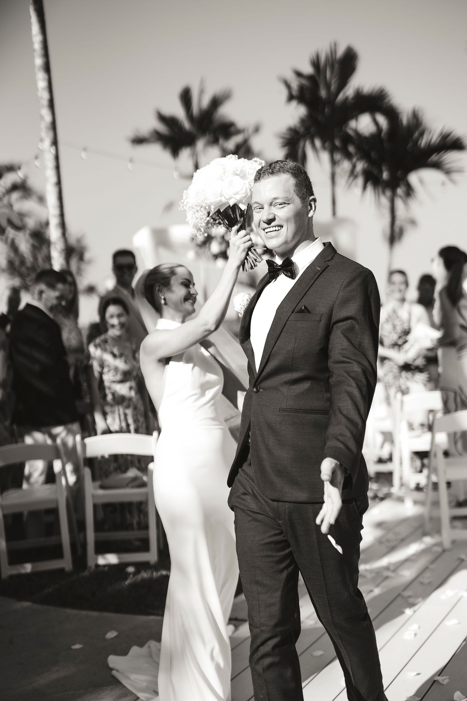 Black and white photo of a smiling groom in a tuxedo walking with his bride in a white gown at an outdoor wedding.