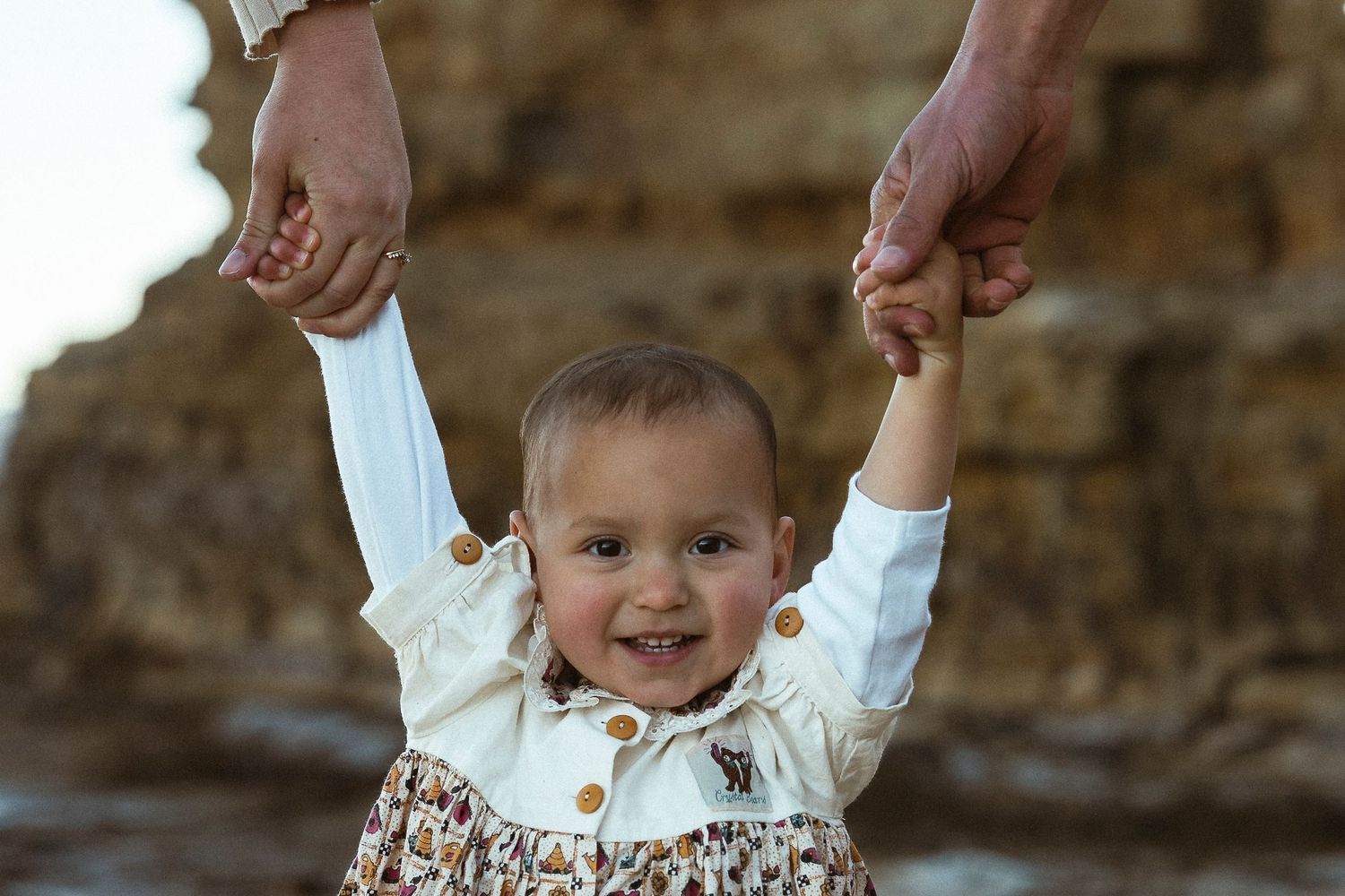 A smiling baby in a floral dress being held up by adult hands outdoors near rocky cliffs.