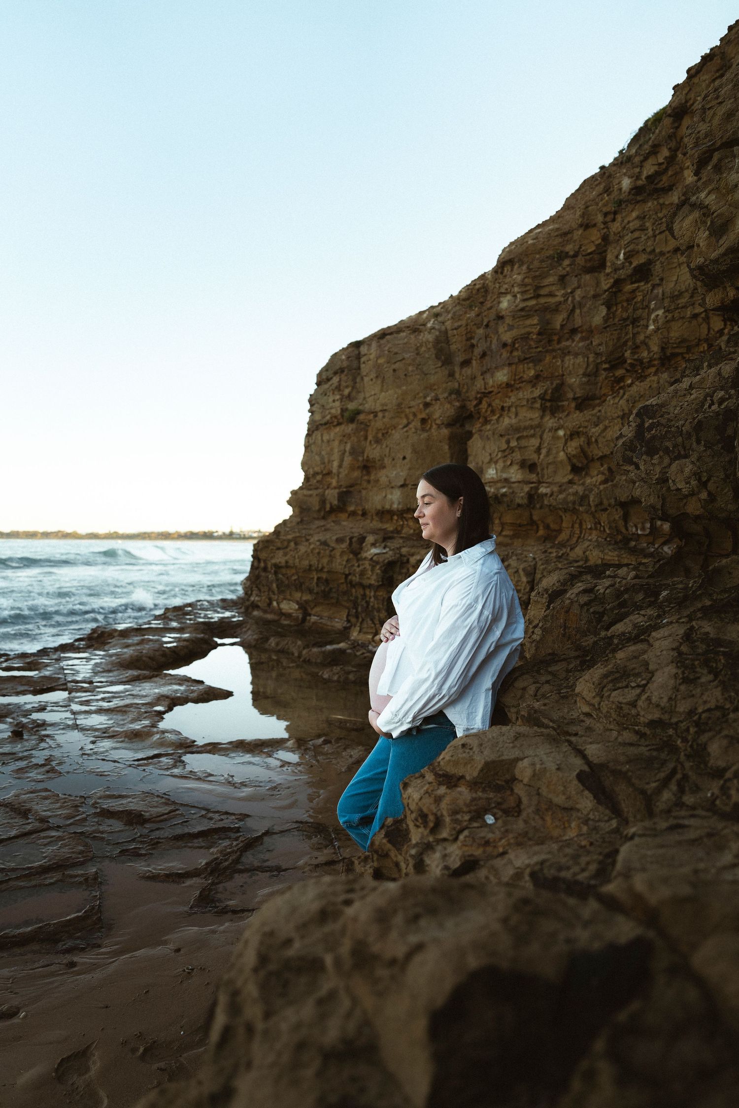 Pregnant woman in white shirt leaning against rocky coastal cliff, eyes closed, embracing her baby bump by the ocean.