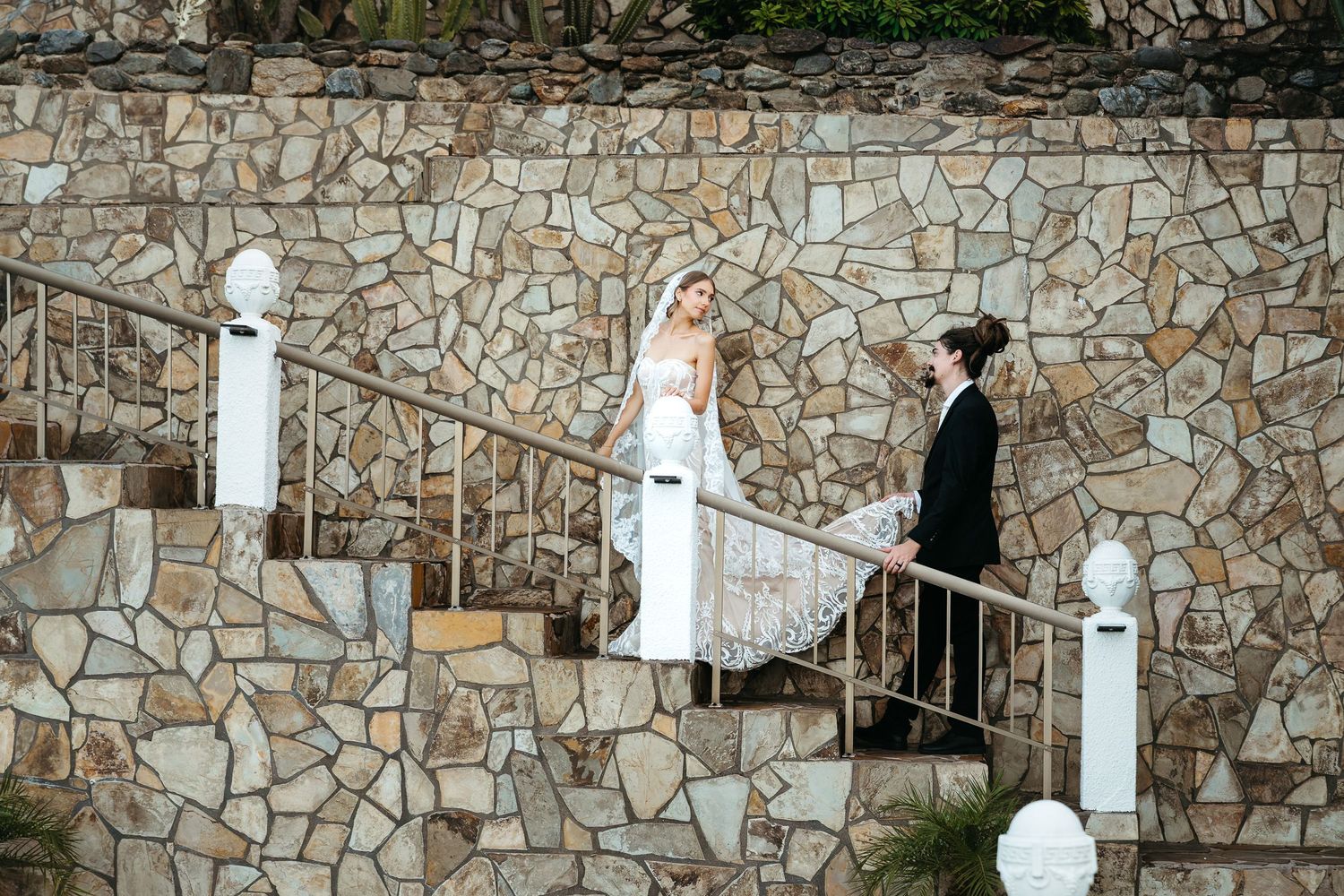 Bride and groom in elegant wedding attire descending stone staircase with metal railing against rustic stone wall backdrop.