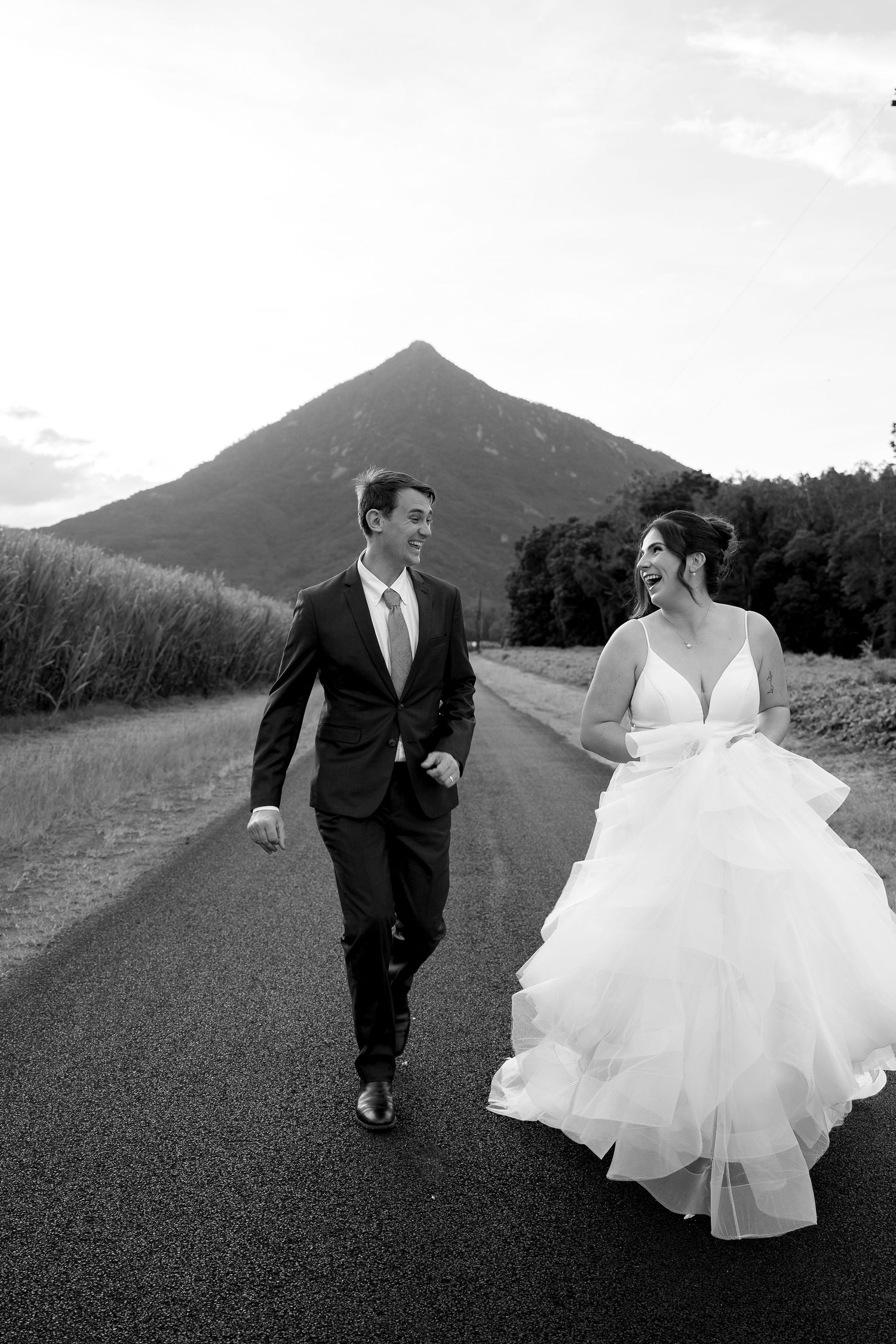Black and white photo of a bride and groom walking and laughing on a road with a mountain backdrop.