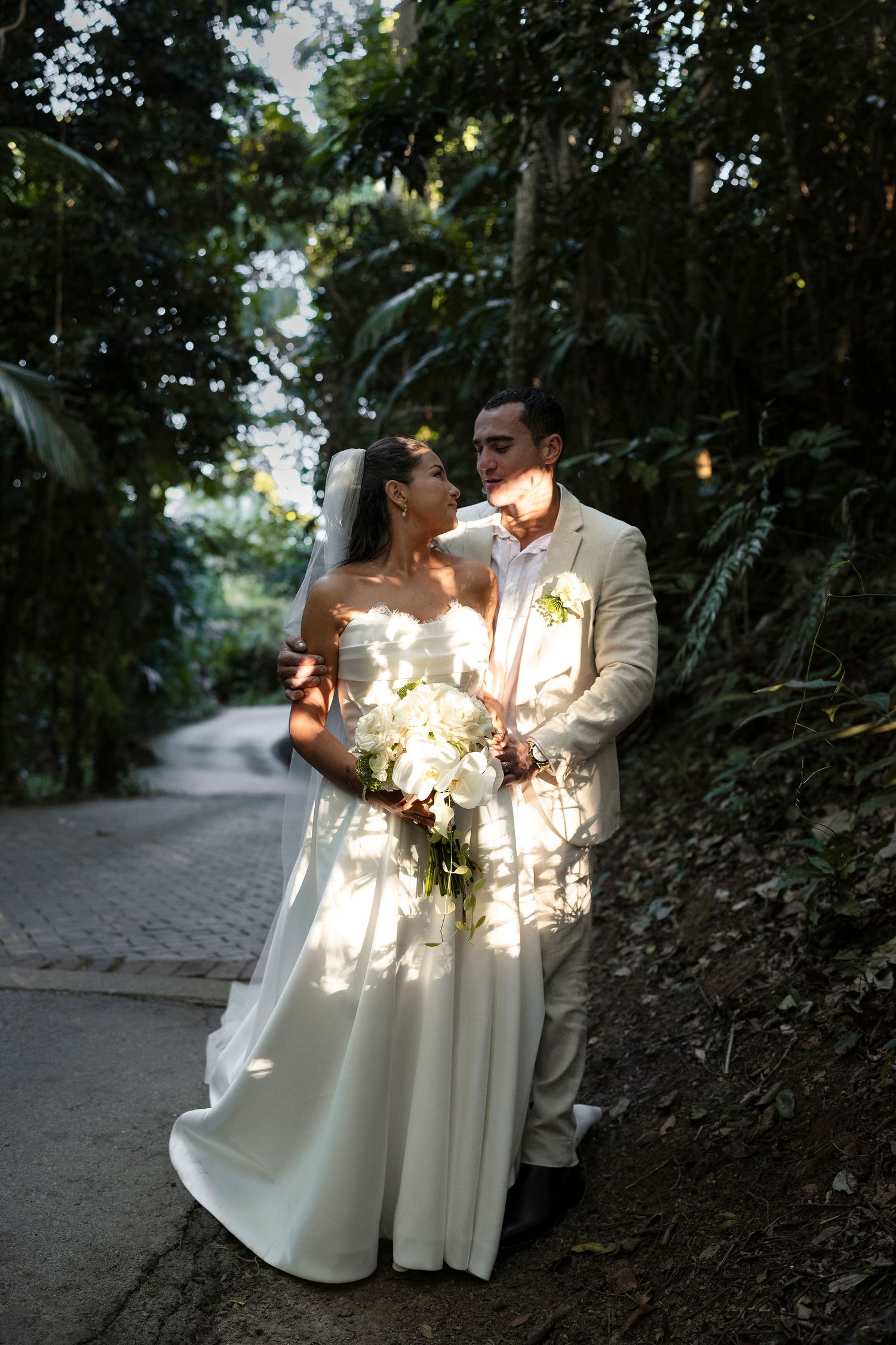 Bride in white gown holding bouquet stands with groom in tan suit against lush tropical greenery in dappled sunlight.