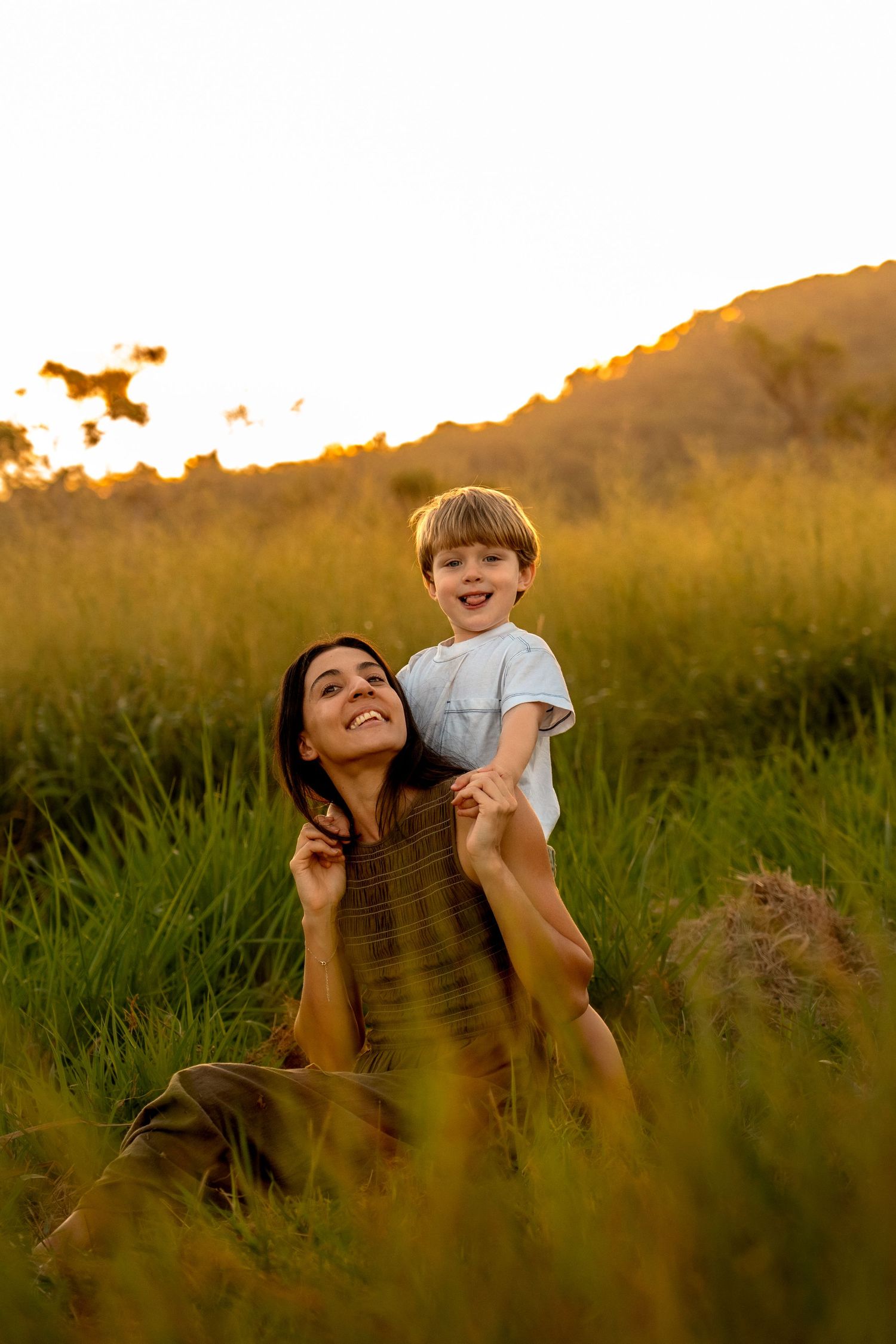 Happy mother sits in golden sunset meadow grass while her laughing young son climbs on her shoulders.