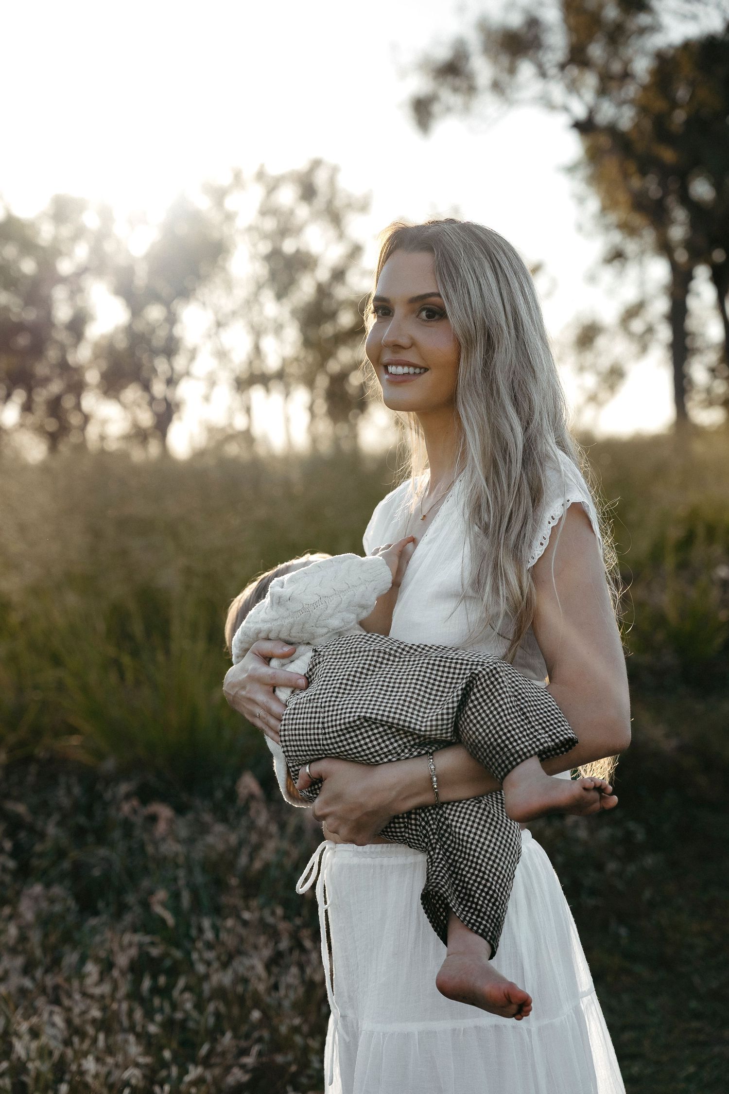 Smiling blonde woman in white dress holding a baby outdoors in golden hour sunlight.
