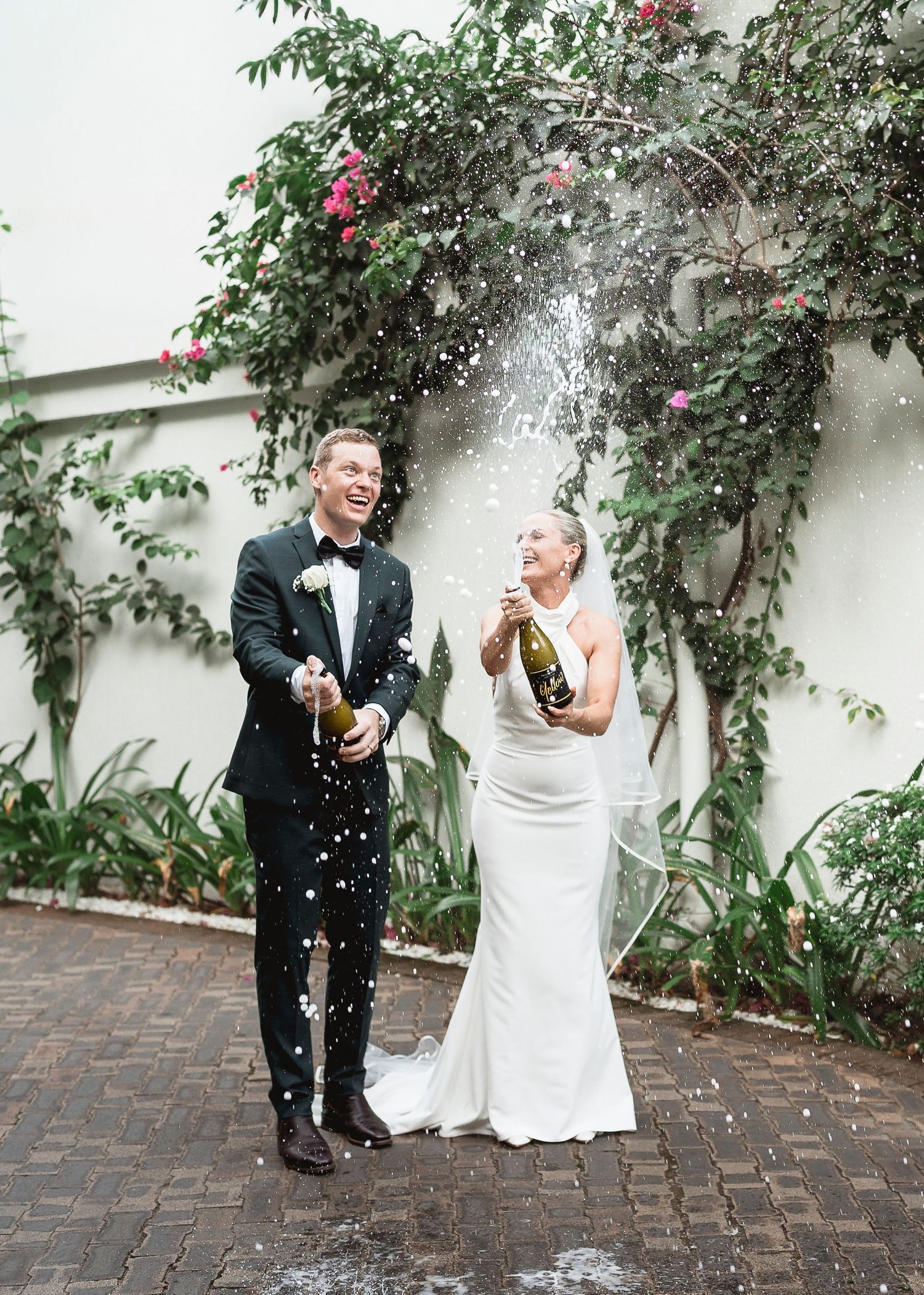 Bride and groom laughing while popping champagne bottles outdoors at their wedding celebration.