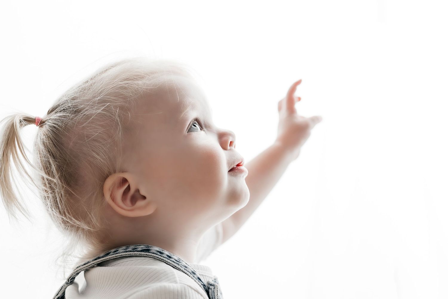 Curious toddler girl with pigtails reaching upward against bright white background.