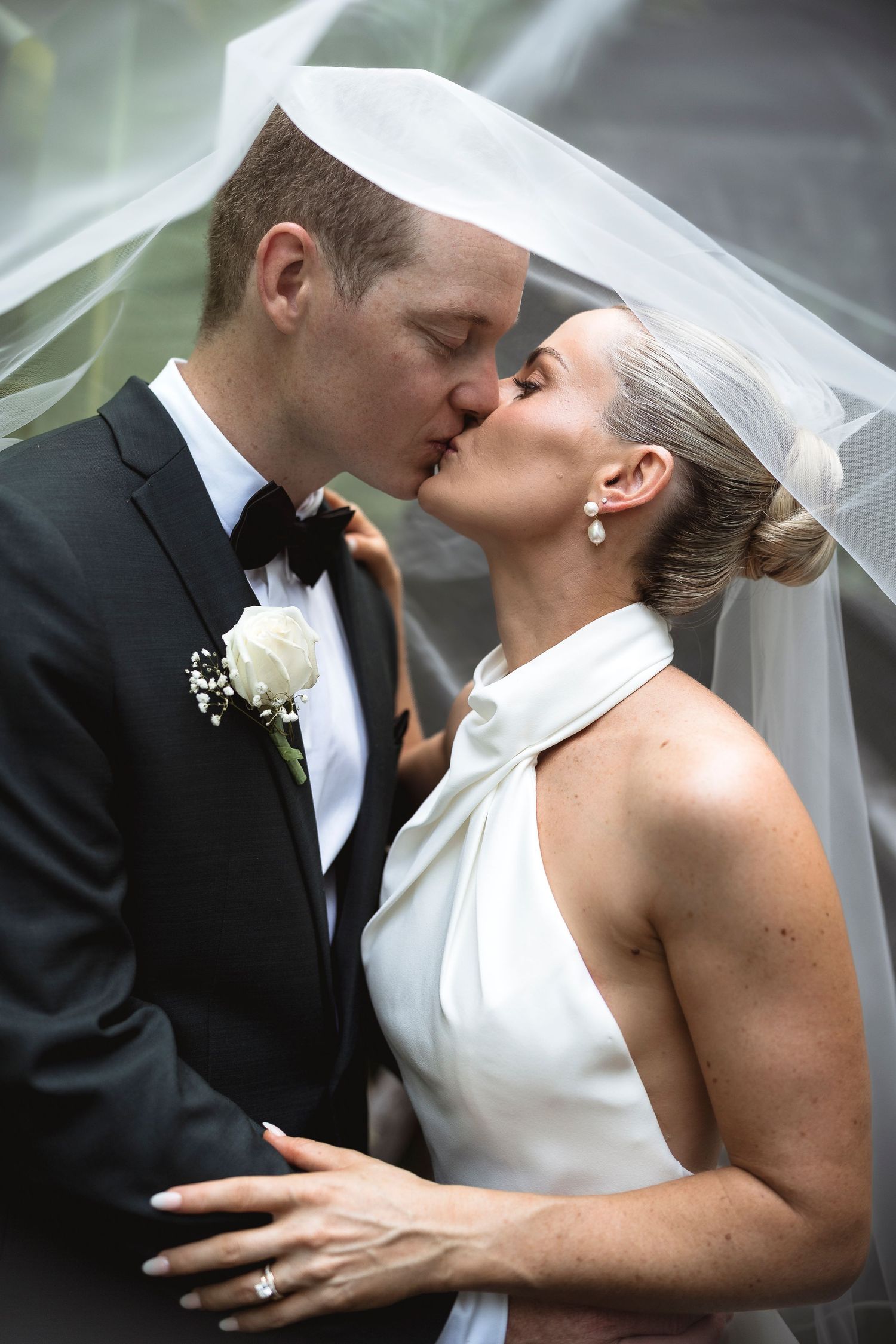 Bride and groom sharing a romantic kiss under a flowing veil, groom in black tuxedo with white rose boutonniere.