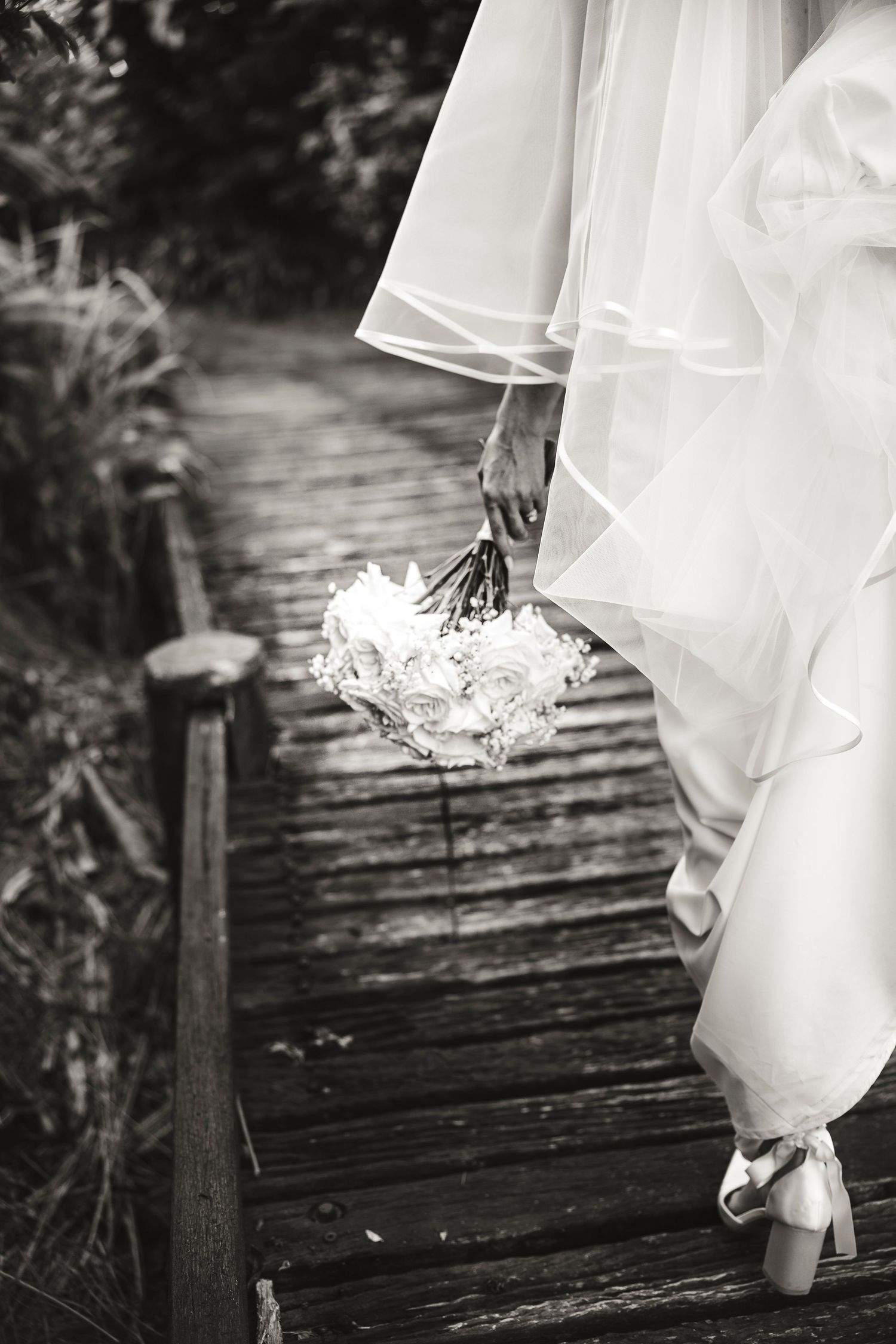 Black and white photo of a bride walking on a wooden bridge, holding a white rose bouquet, wearing a flowing gown.