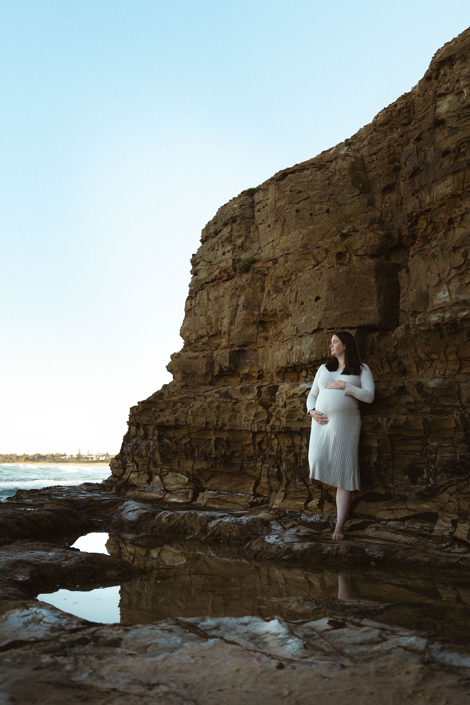 Pregnant woman in white dress leaning against rocky coastal cliffs, cradling her baby bump by ocean tide pools.