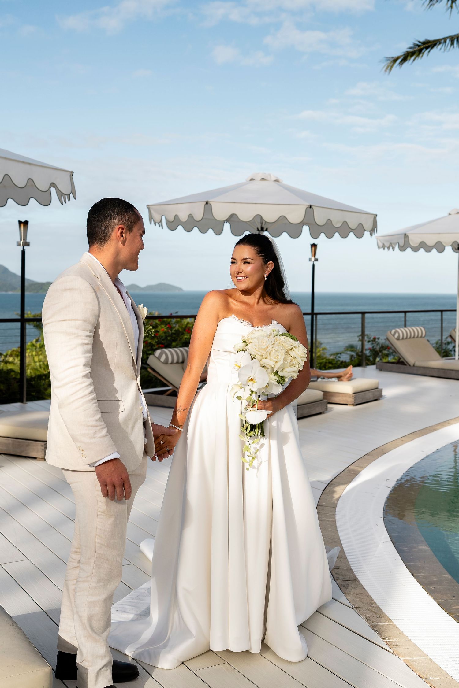 Bride and groom in white wedding attire stand by a poolside terrace with ocean views and elegant umbrellas.
