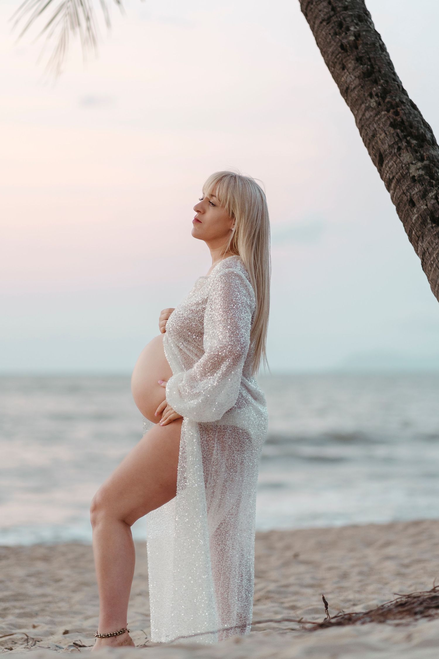 Pregnant woman in sheer white dress posing on tropical beach at sunset near palm tree.