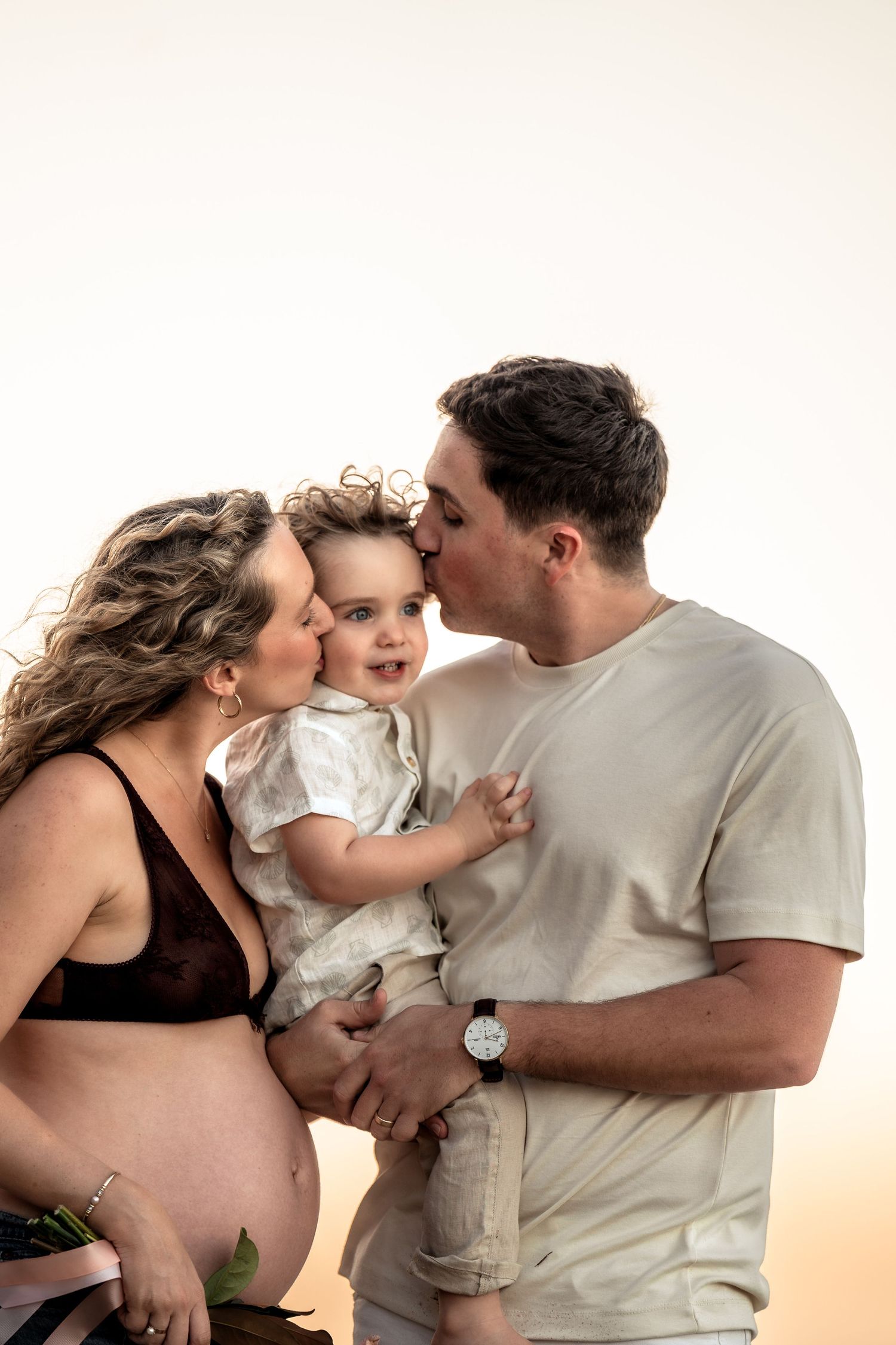 Couple kissing their smiling toddler on the cheeks during a maternity photo session.