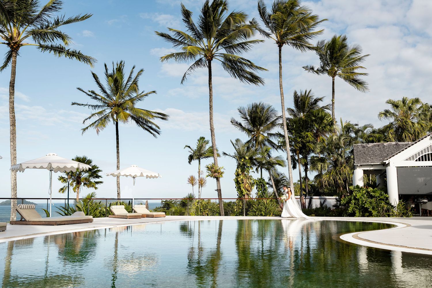 Bride and groom pose by a tropical resort infinity pool surrounded by tall palm trees with ocean views.