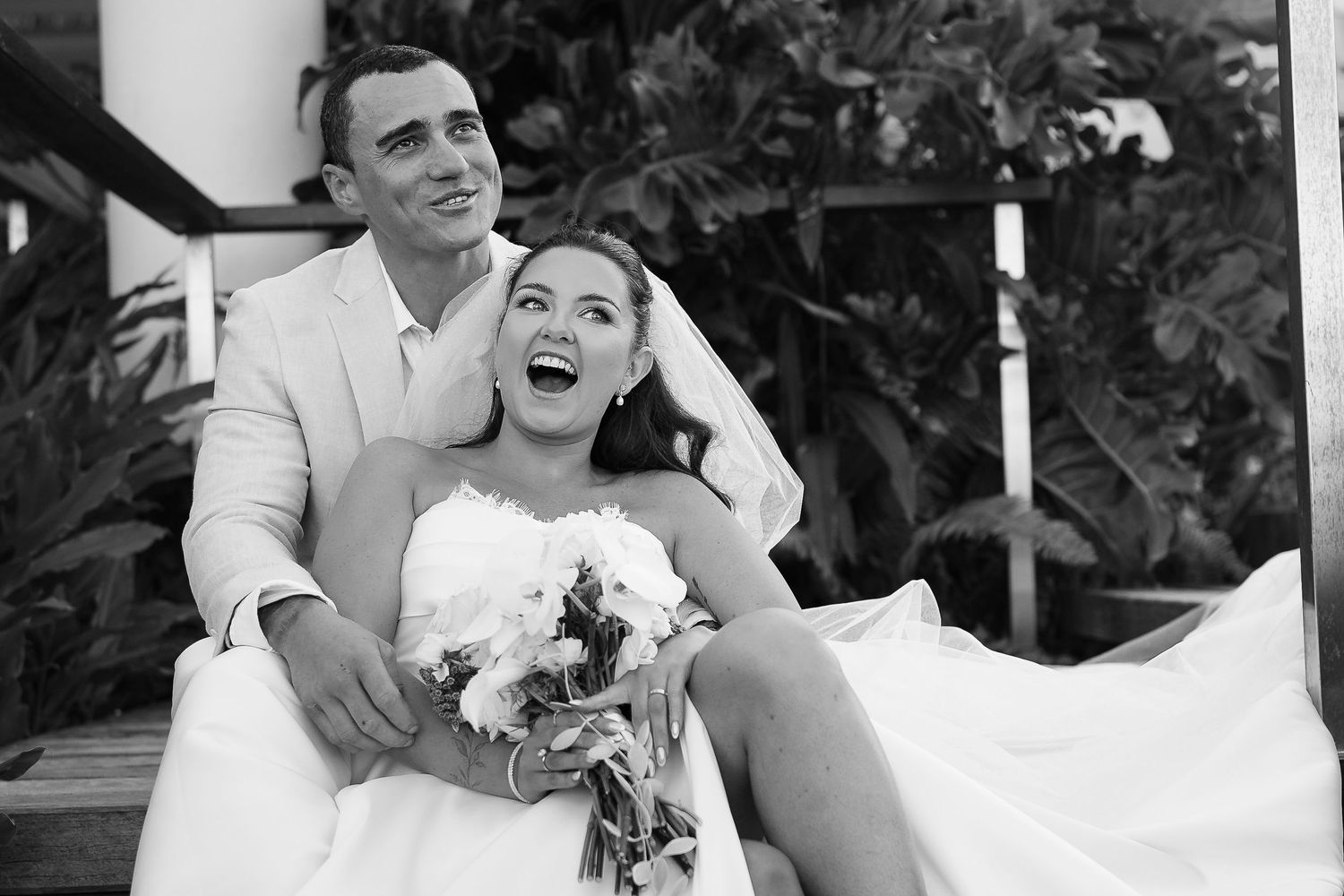 Black and white photo of a joyful bride and groom sitting together outdoors, bride holding white bouquet.