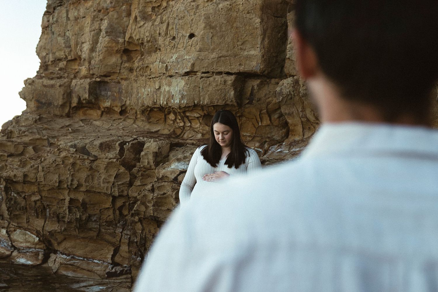 Couple standing near rocky coastal cliffs, woman in white dress looking down, man in foreground out of focus.