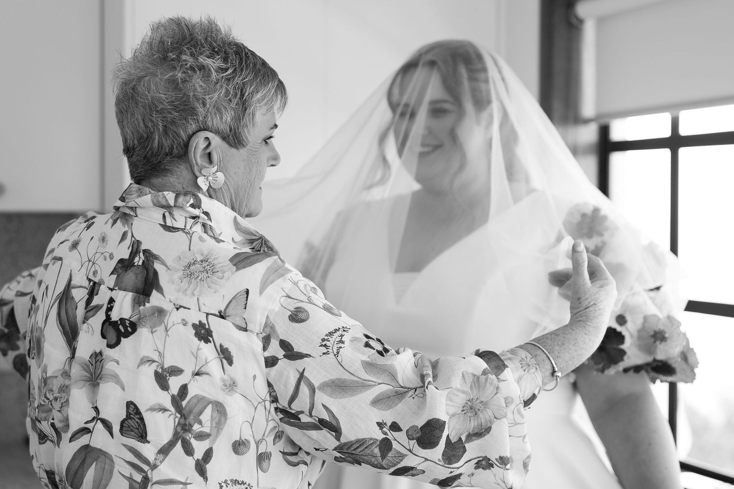 Black and white photo of a bride in veil being helped by an older woman in a floral blouse on her wedding day.