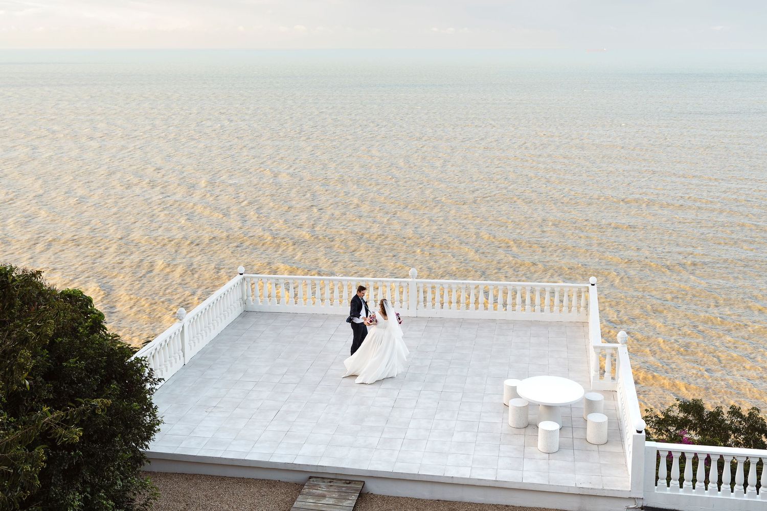 Bride and groom dance on a white terrace overlooking a calm ocean at sunset during their beachside wedding.