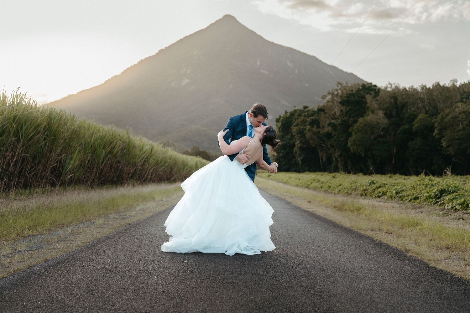 Bride and groom share romantic dip kiss on open road with scenic mountain backdrop at sunset.