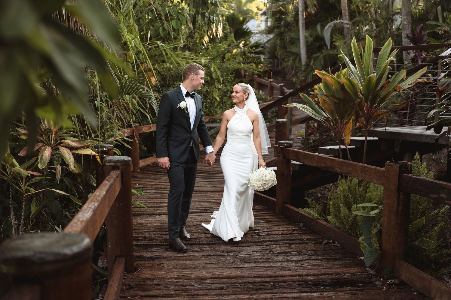 Bride and groom holding hands on a wooden bridge surrounded by lush tropical greenery on their wedding day.
