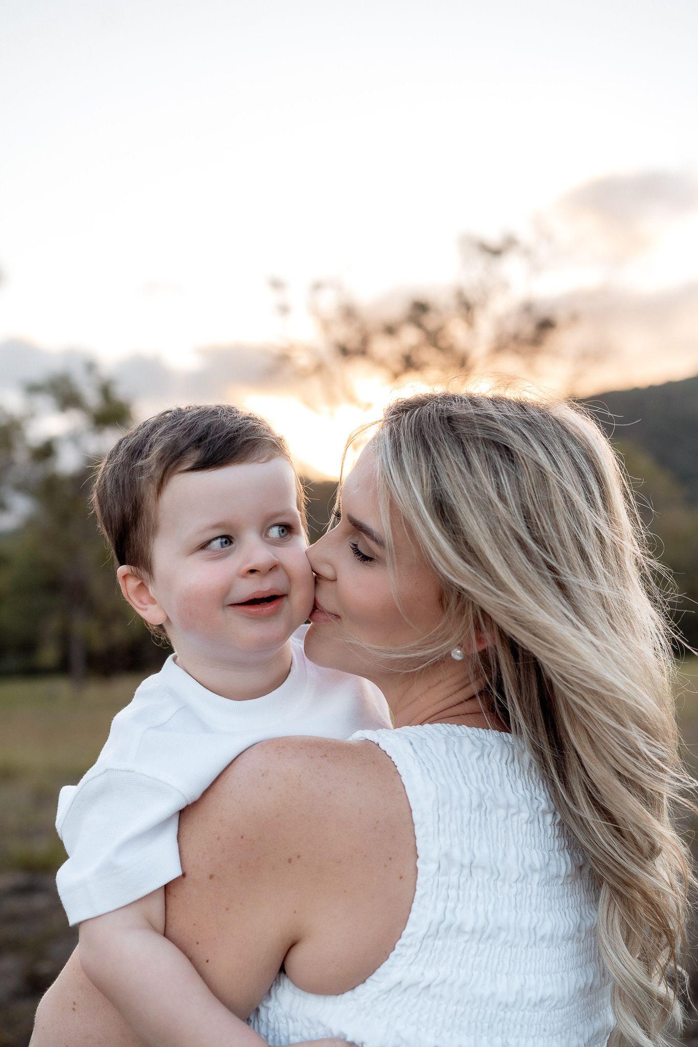 Blonde mother kissing smiling toddler on the cheek at golden hour sunset outdoors.