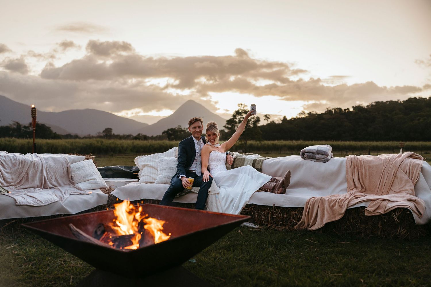 Bride and groom celebrate by a fire pit at sunset with mountain views and white draped lounge furniture.