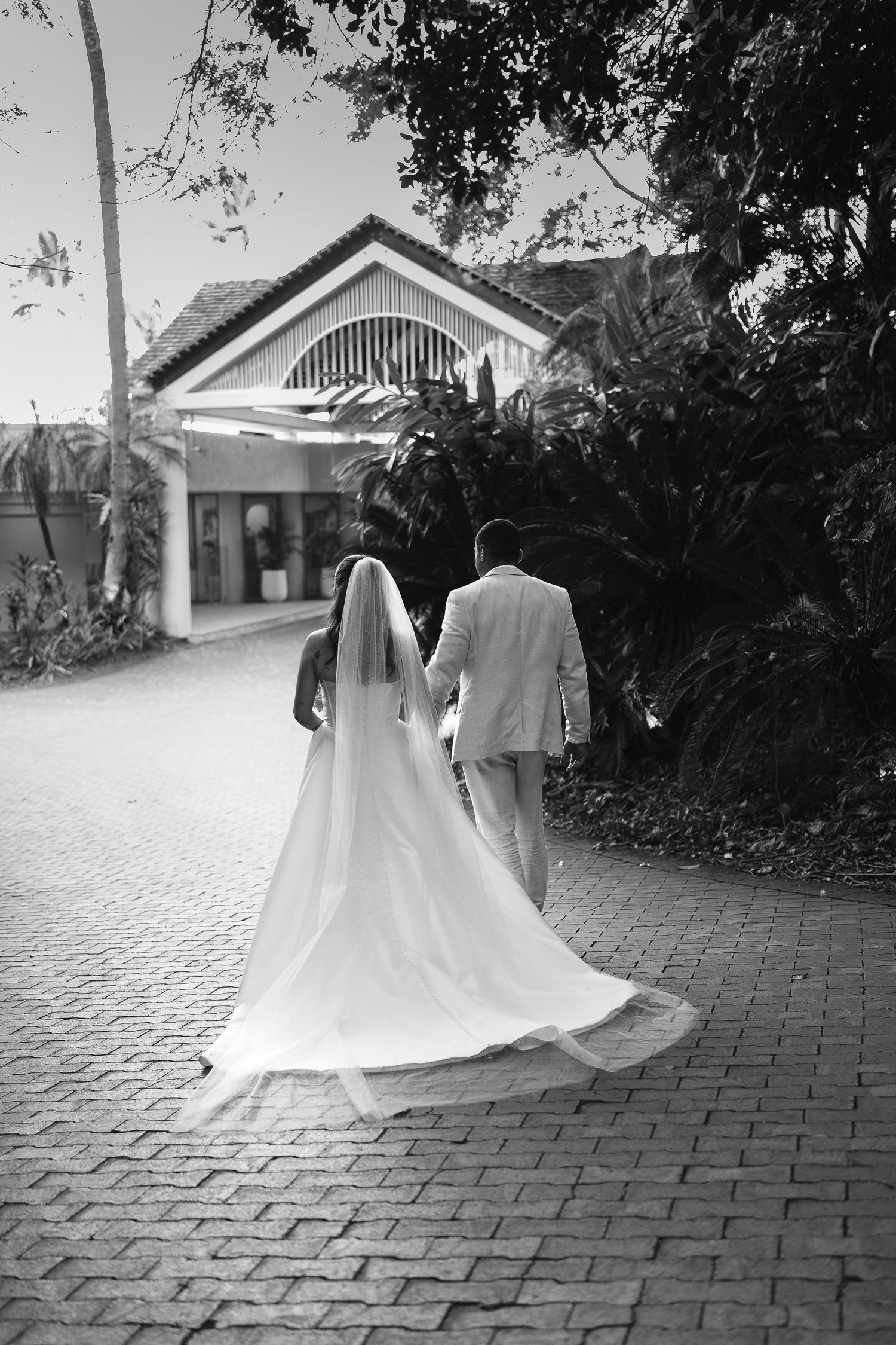 Black and white photo of bride and groom walking hand in hand toward a building surrounded by lush tropical greenery.