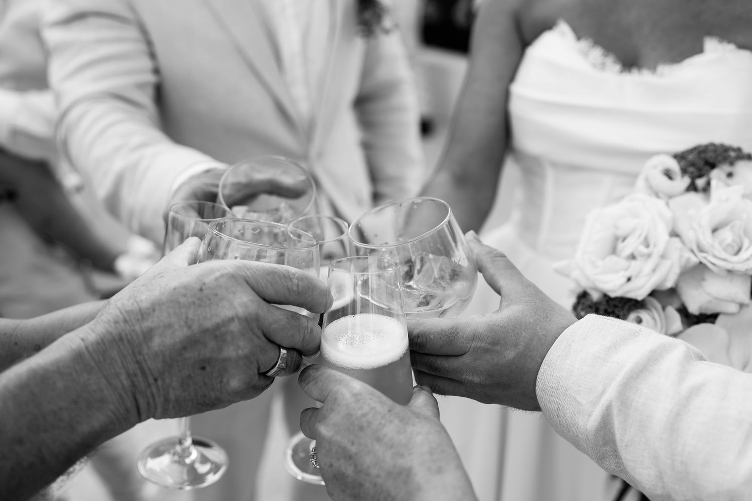 Black and white photo of wedding guests clinking champagne glasses in a celebratory toast, with bride holding bouquet.