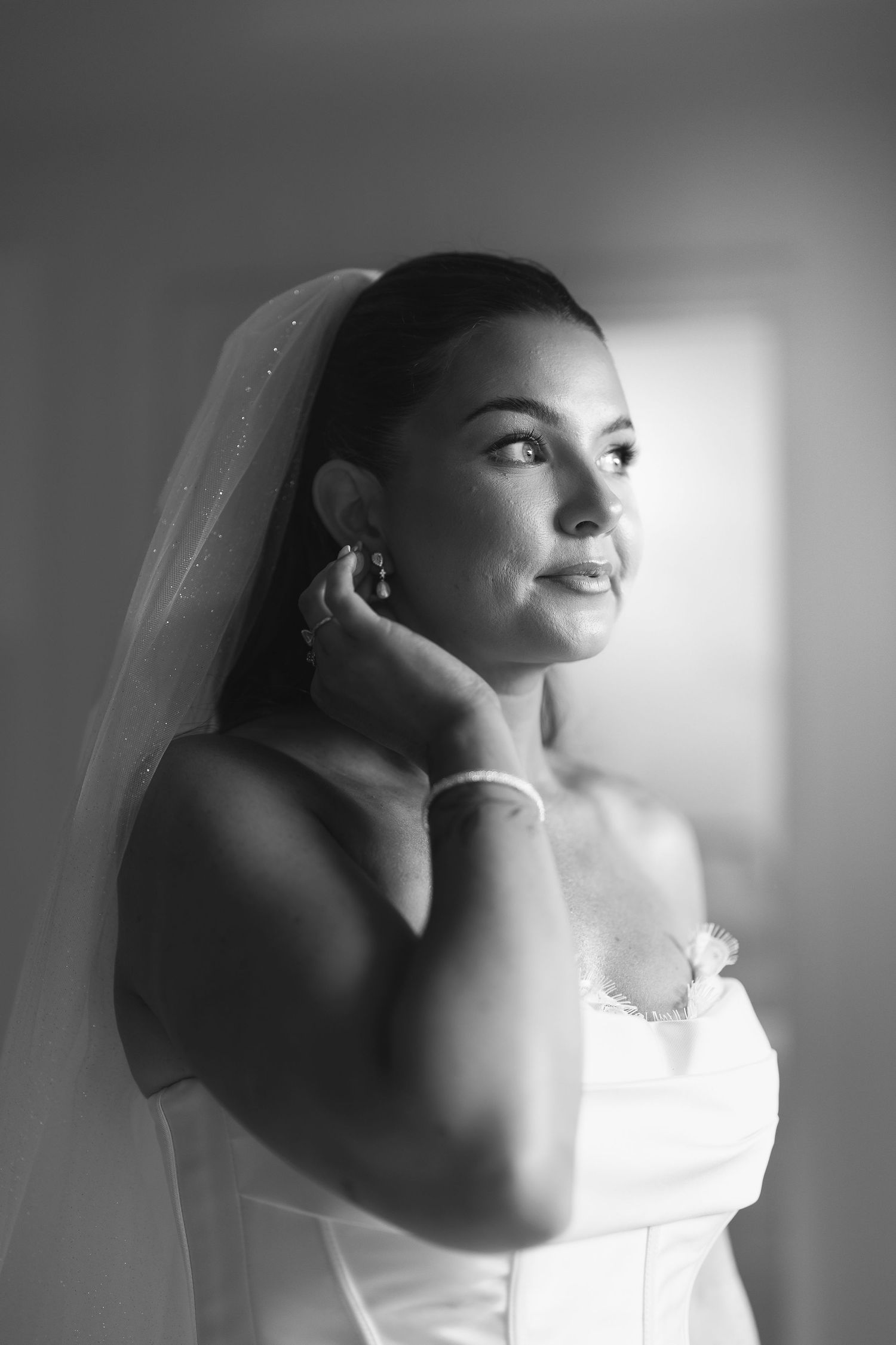 Black and white portrait of a bride in a strapless gown and veil, touching her earring while gazing softly to the side.