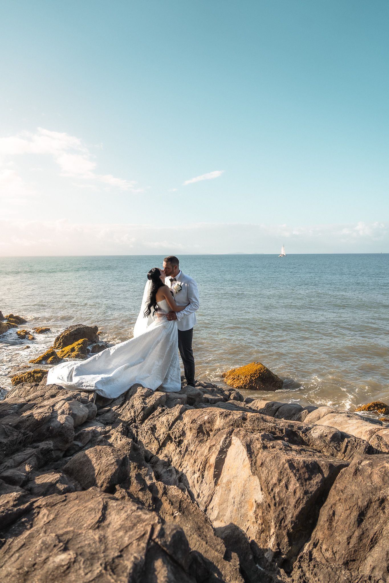 Bride and groom embracing on rocky shoreline with ocean and blue sky backdrop.