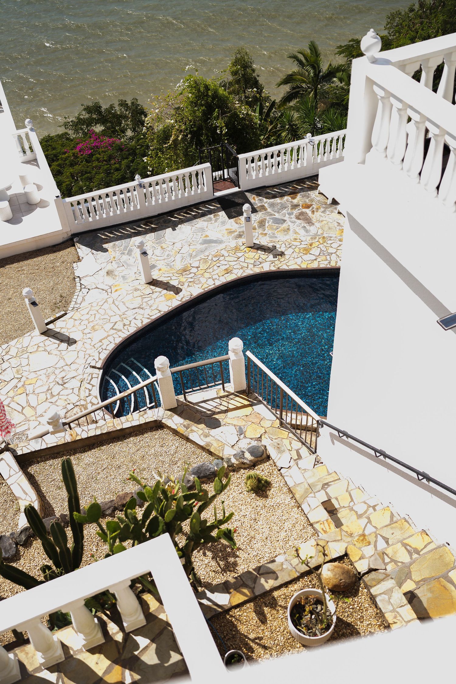 Aerial view of a luxury villa with a blue swimming pool, mosaic stone patio, white balustrades, and ocean backdrop.