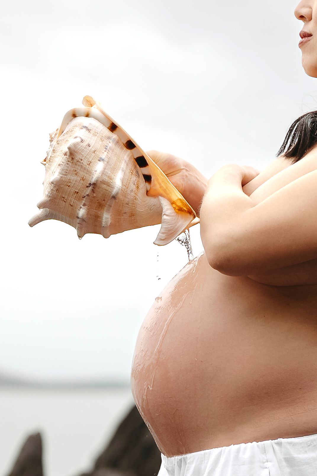 Pregnant woman holding a large conch shell with water dripping onto her bare belly outdoors.