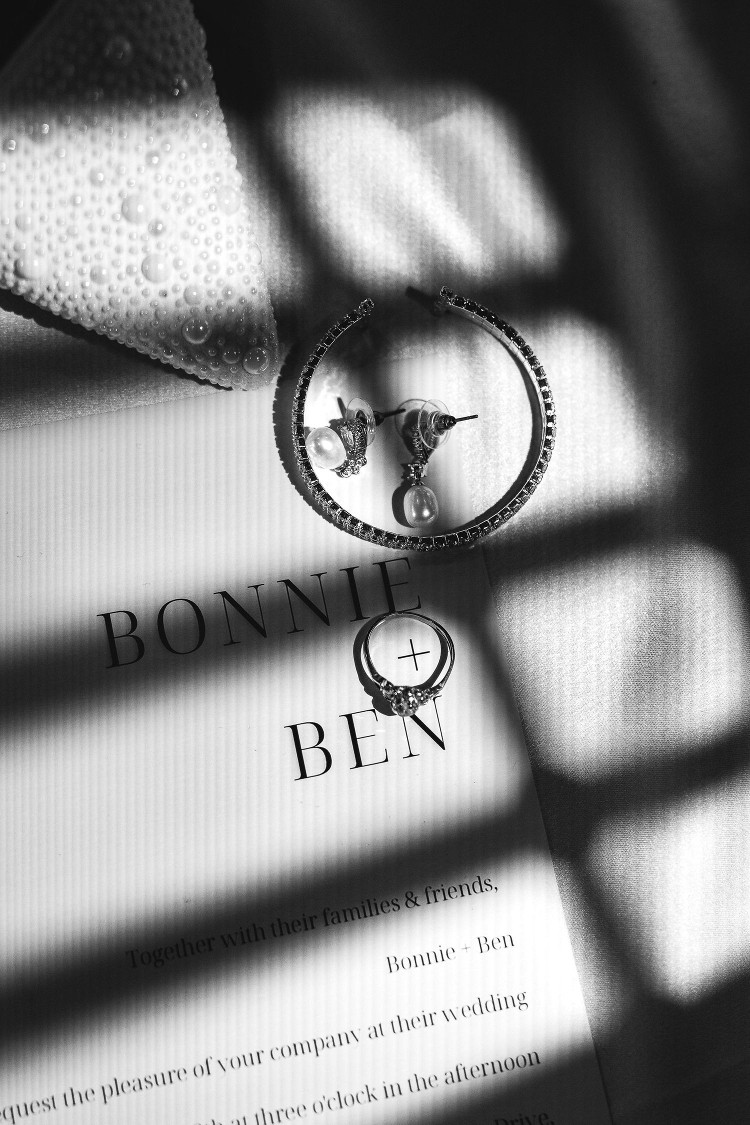 Black and white photo of hoop earrings on a Bonnie and Ben wedding invitation with dramatic shadow overlay.