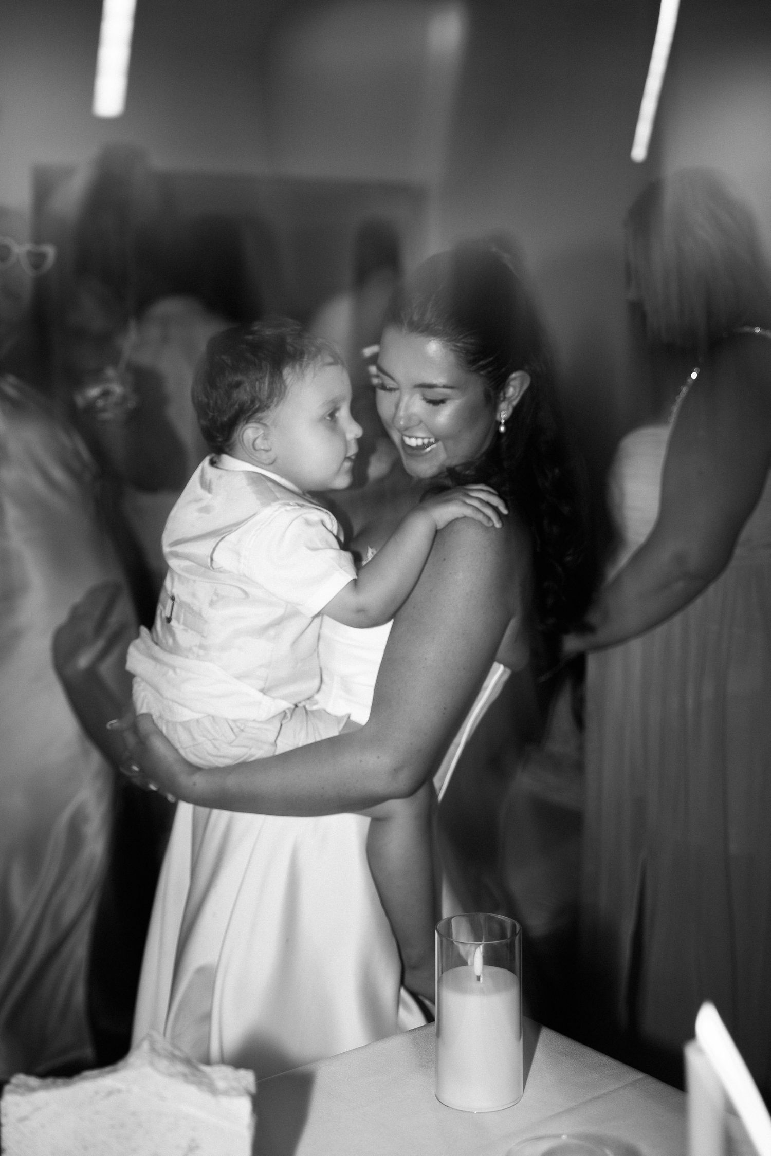 Black and white photo of a bride in white dress holding a baby at a wedding reception with candles.