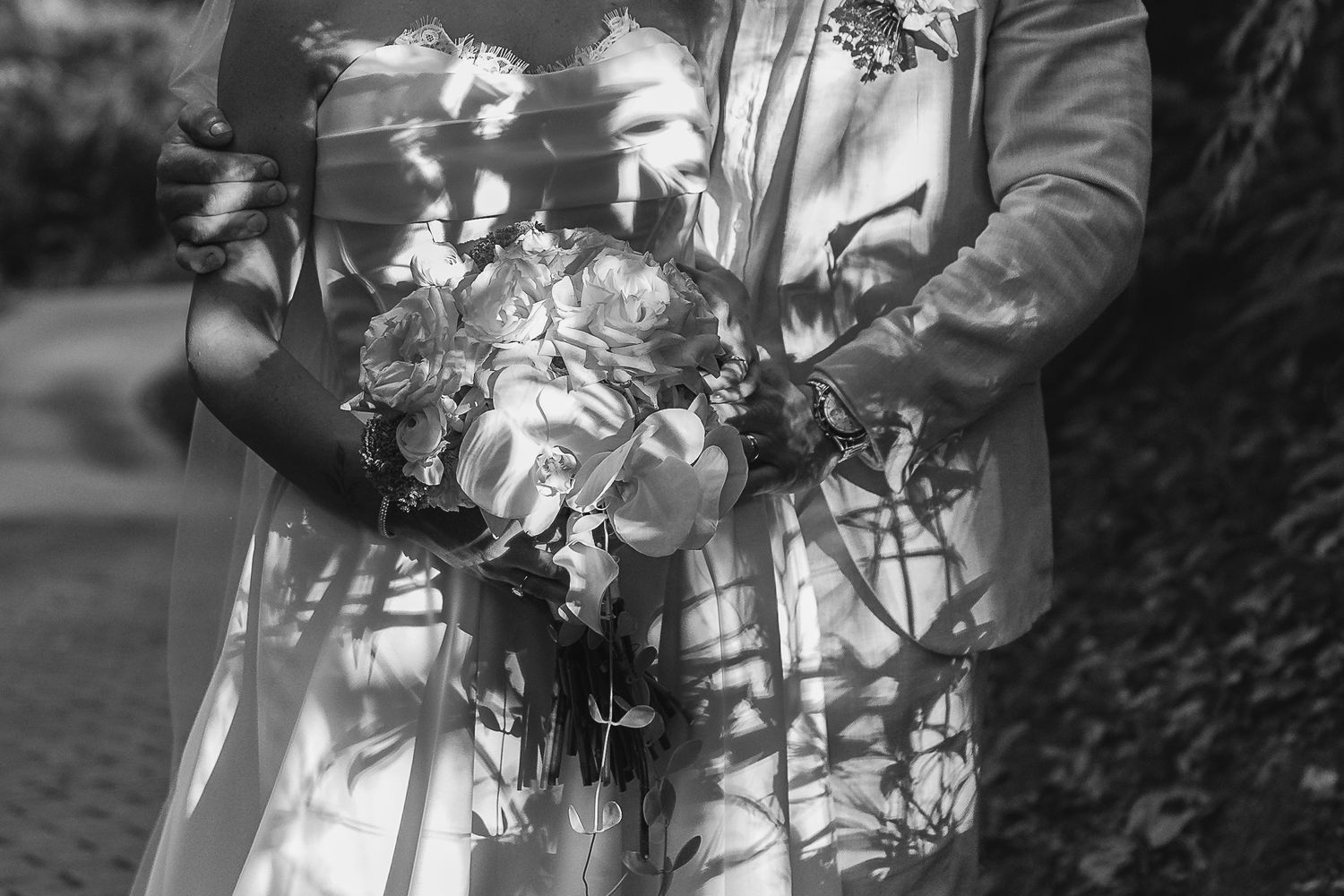 Black and white photo of a bride and groom holding a floral bouquet, with dappled sunlight casting shadows.