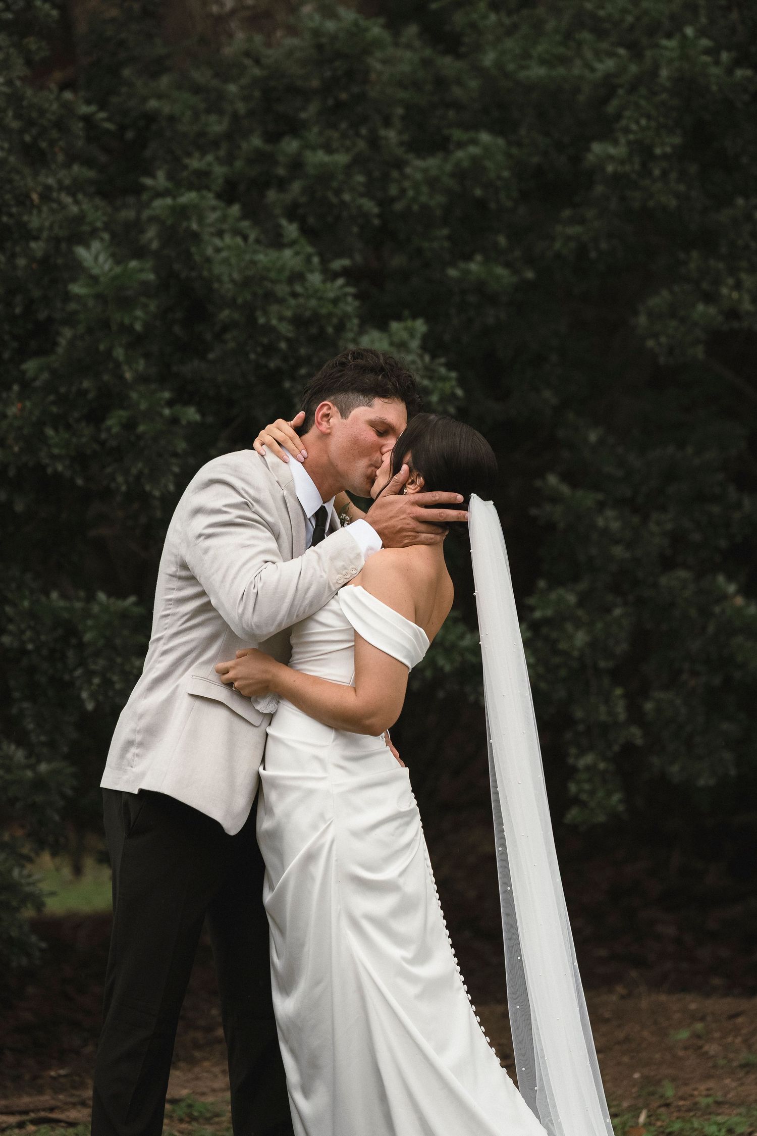 A bride and groom share a romantic kiss outdoors, surrounded by lush green trees, as she wears a white gown and veil.