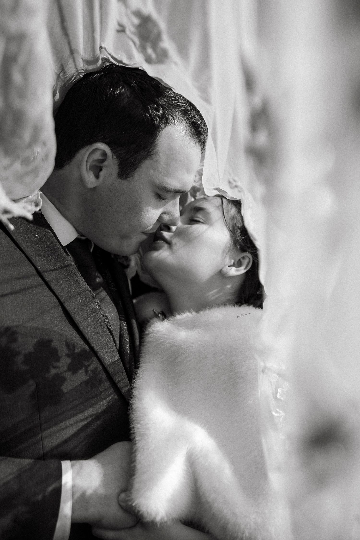 Black and white photo of a bride and groom sharing a romantic kiss, bride wearing a fur wrap and veil.