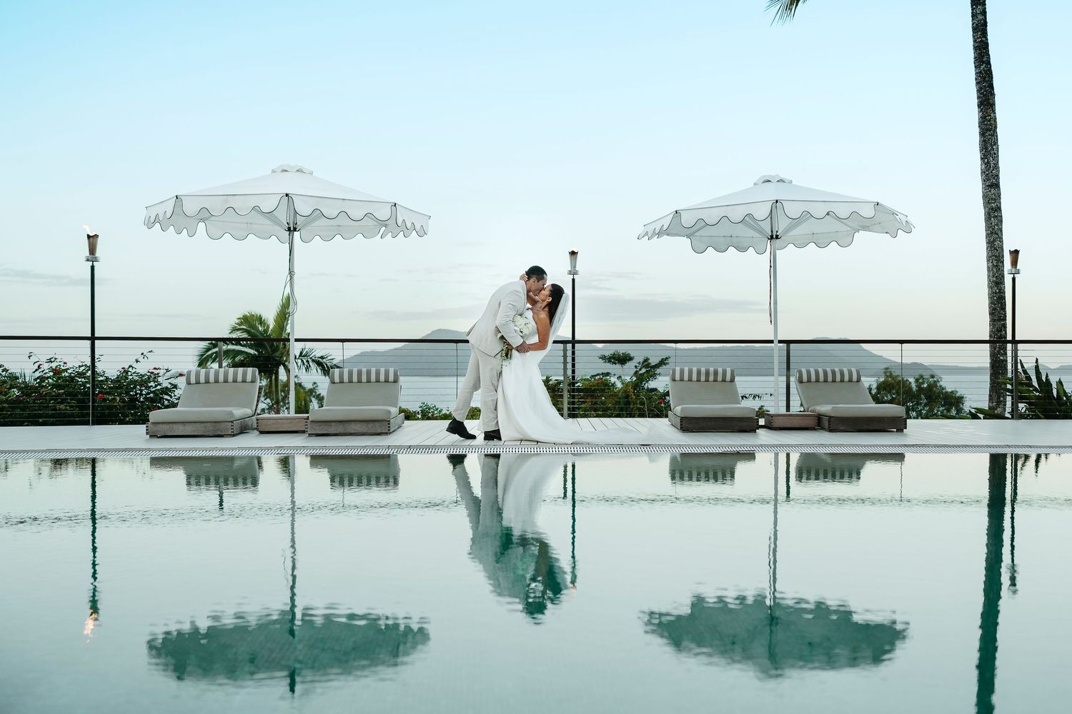 Bride and groom kissing poolside at a luxury resort with white umbrellas and ocean views on their wedding day.
