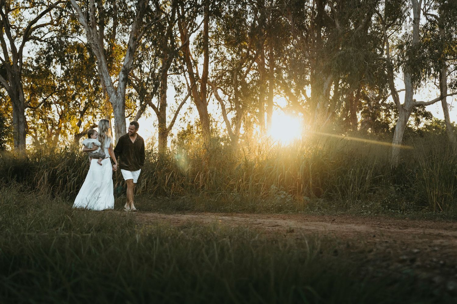 Couple in wedding attire walking through sunlit forest at golden hour, sun rays filtering through trees.