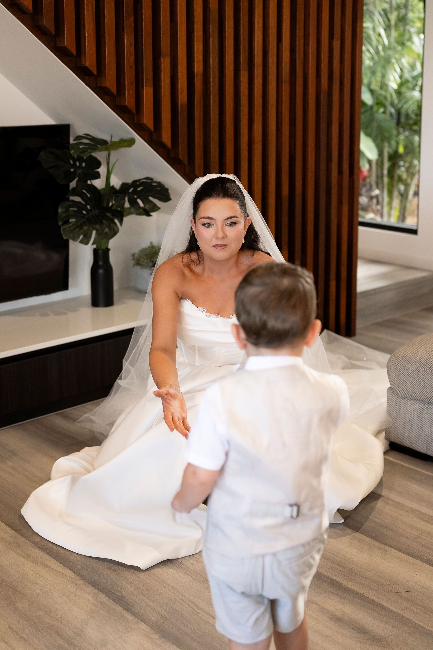 Bride in white wedding gown and veil kneeling to greet a young boy in white shirt indoors.