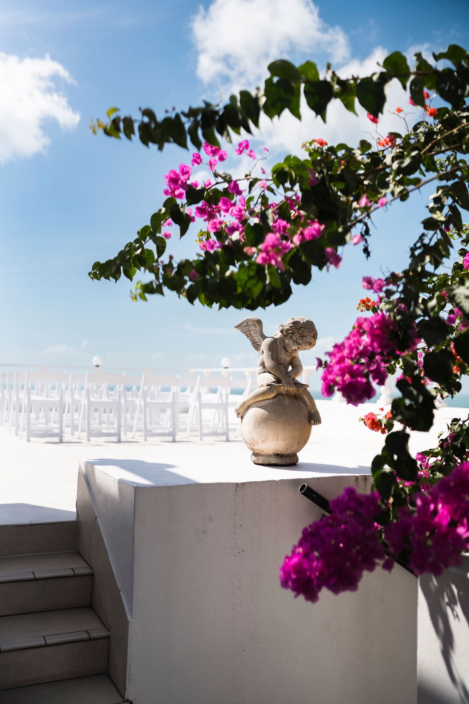 White wedding chairs set outdoors near bougainvillea flowers and angel statue under blue sky.