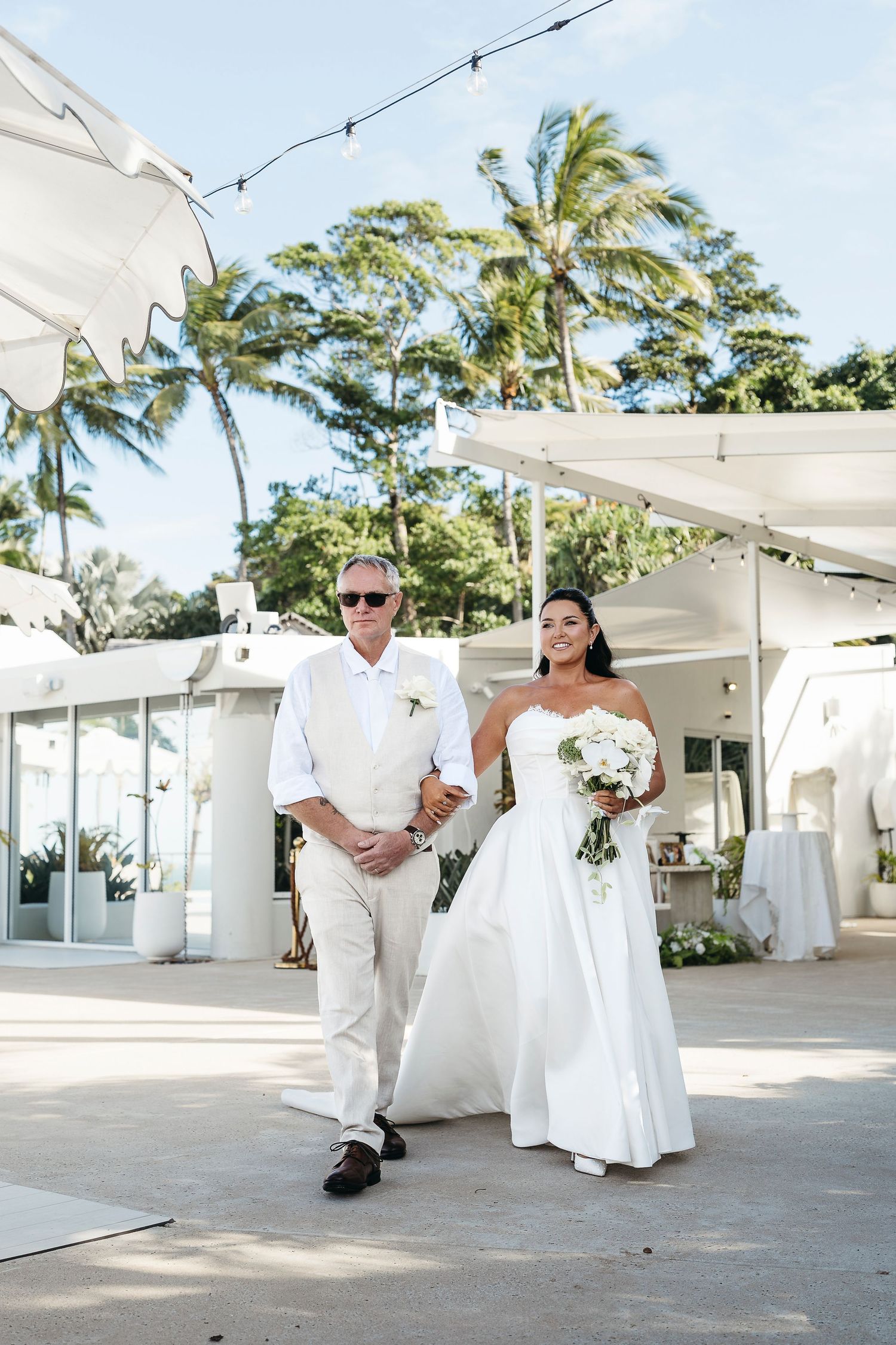 Bride in strapless white gown walks with older man in white suit at outdoor tropical wedding venue.