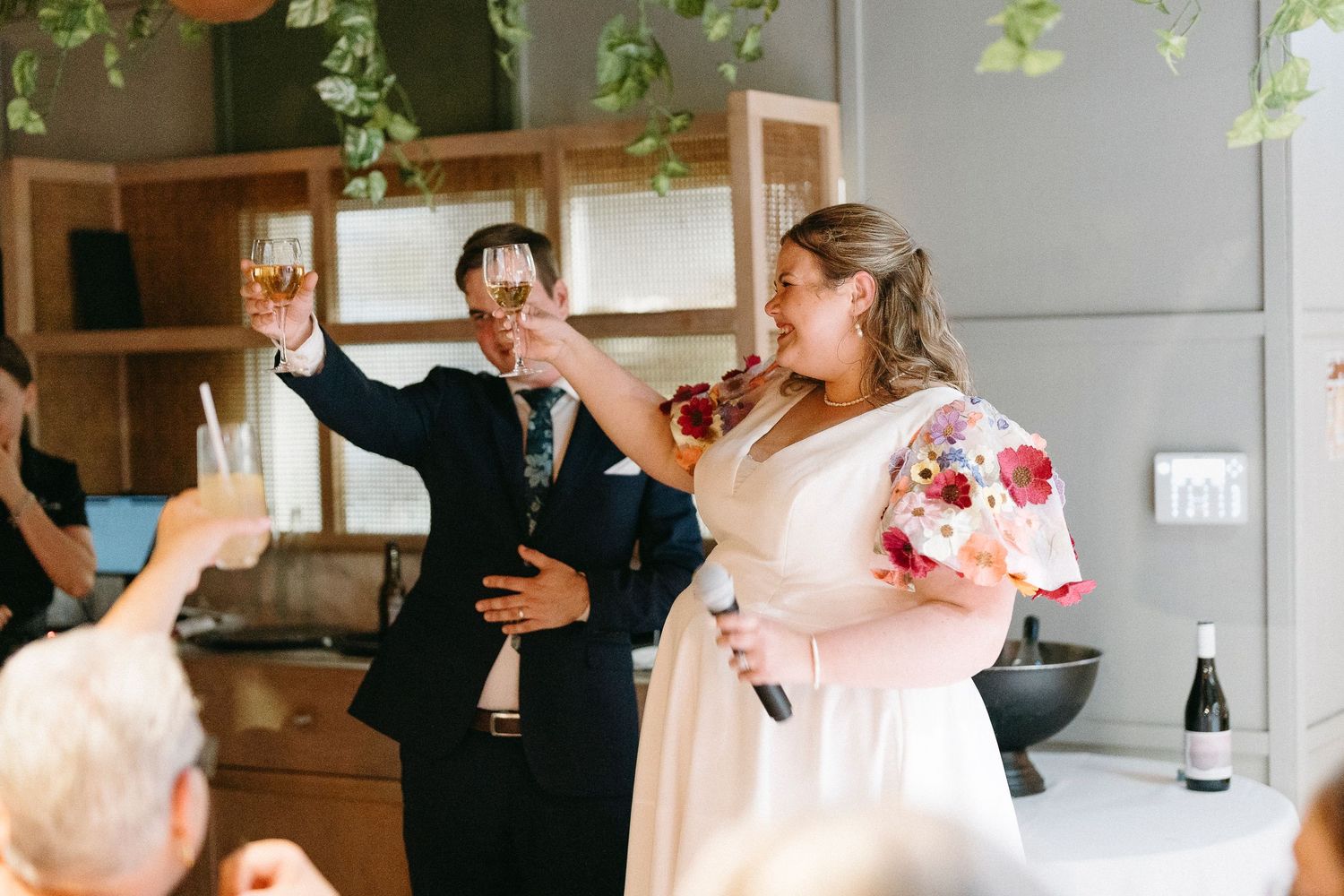 Bride and groom toasting with champagne glasses at wedding reception, bride holding colorful floral bouquet.