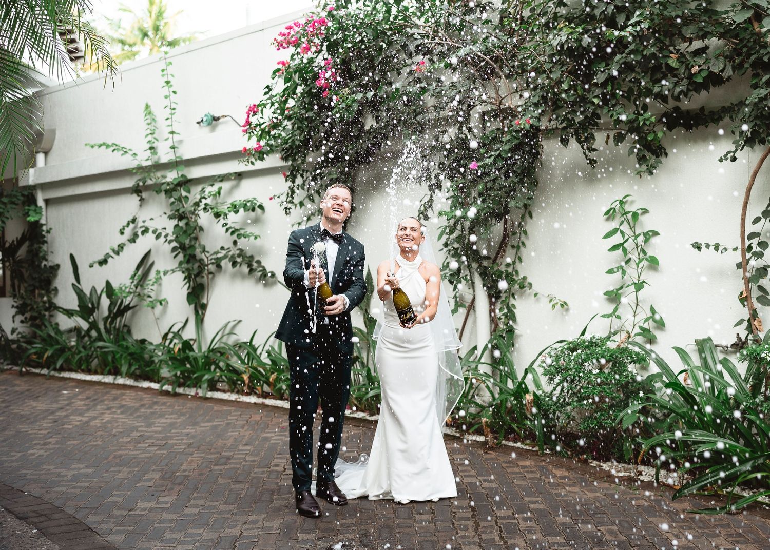 Joyful newlyweds popping champagne bottles outdoors, surrounded by lush greenery and falling confetti on their wedding day.