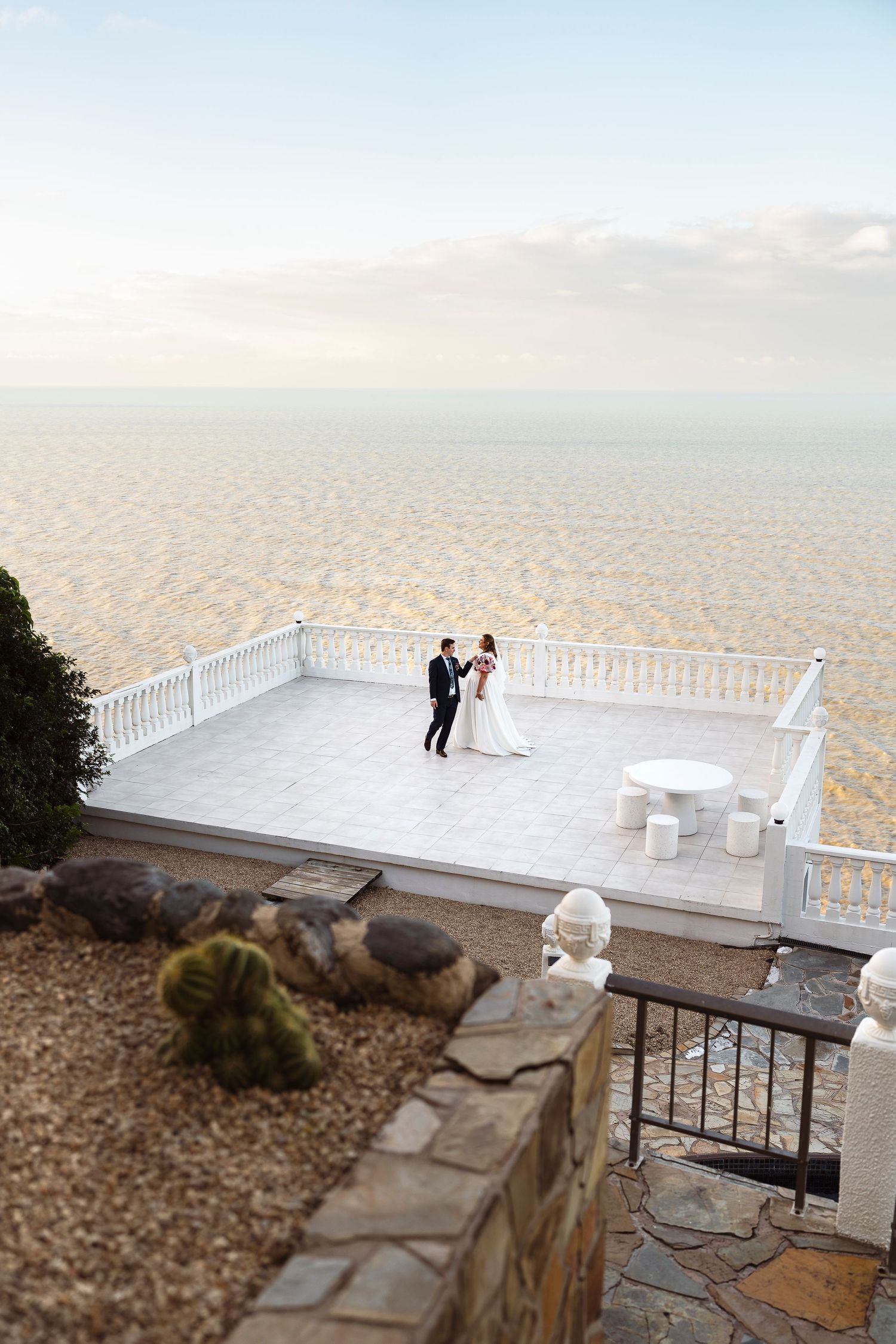 Bride and groom share a romantic moment on a white oceanfront terrace balcony overlooking a vast sandy beach.