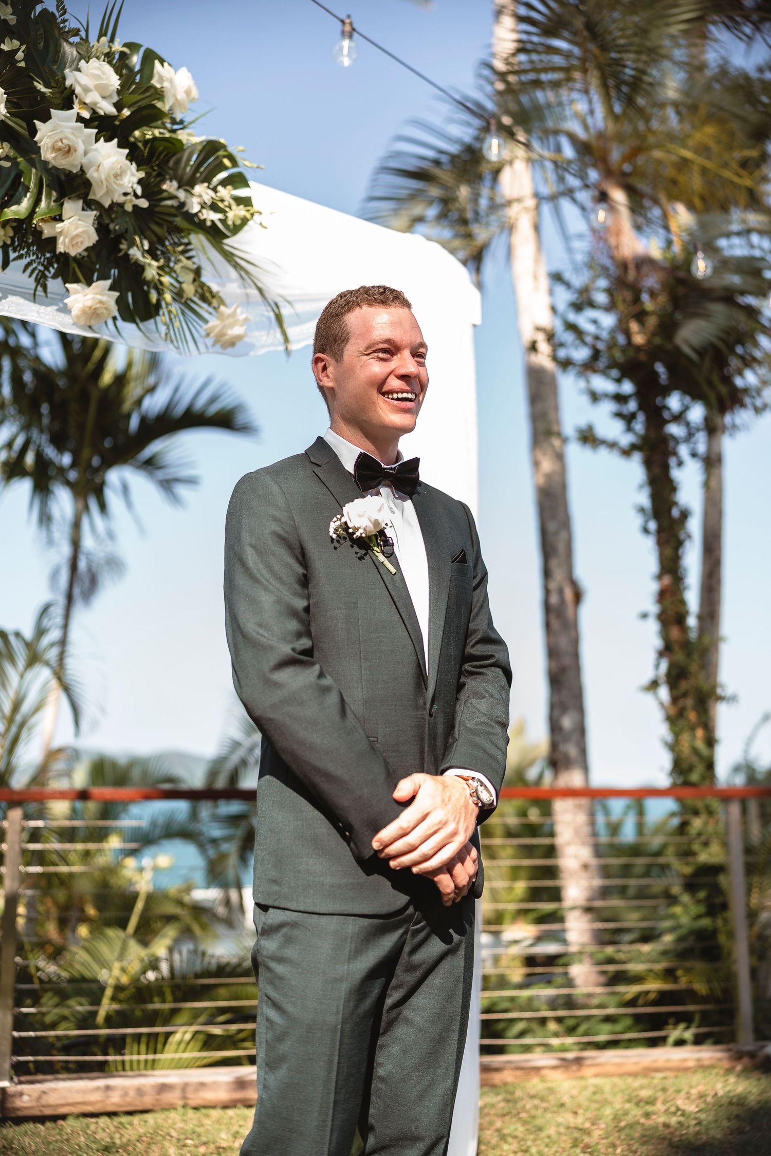 Smiling groom in gray tuxedo with boutonniere stands at outdoor wedding altar with white arch and tropical palms.