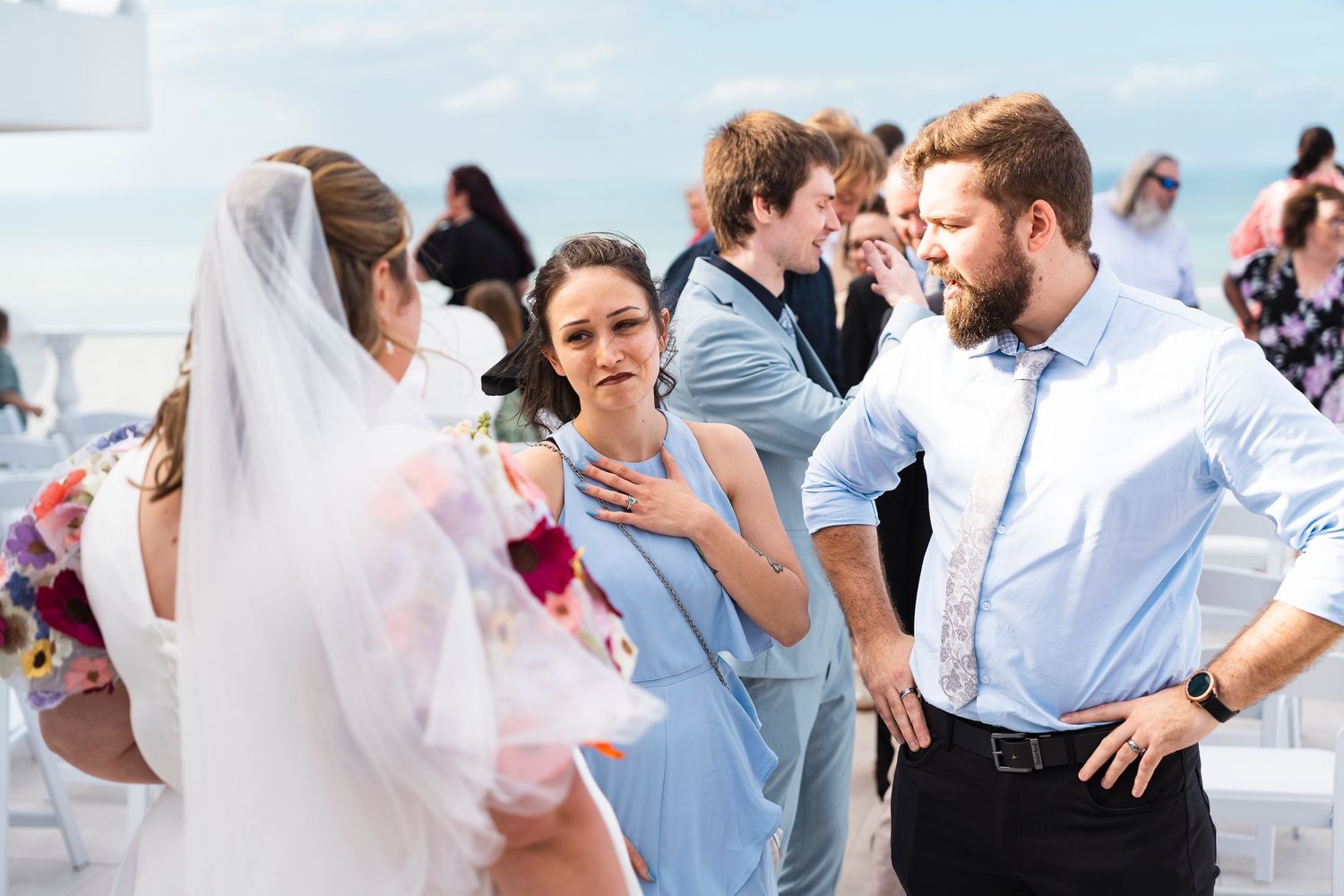 Bride in white veil speaks with guests in blue attire at an outdoor beach wedding ceremony.