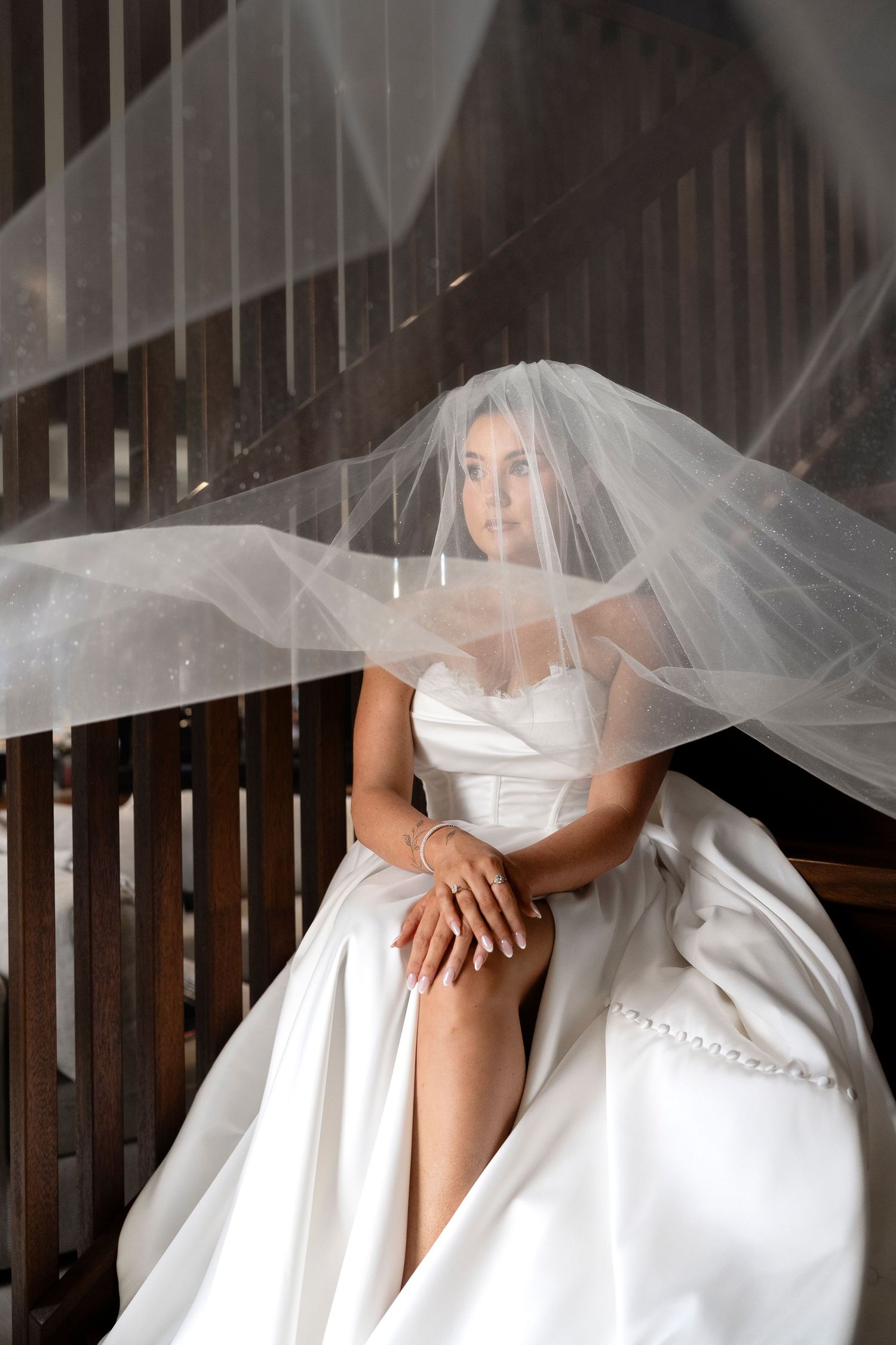 Elegant bride in white strapless gown and flowing veil sits gracefully, showcasing bridal jewelry and pearl-adorned dress.