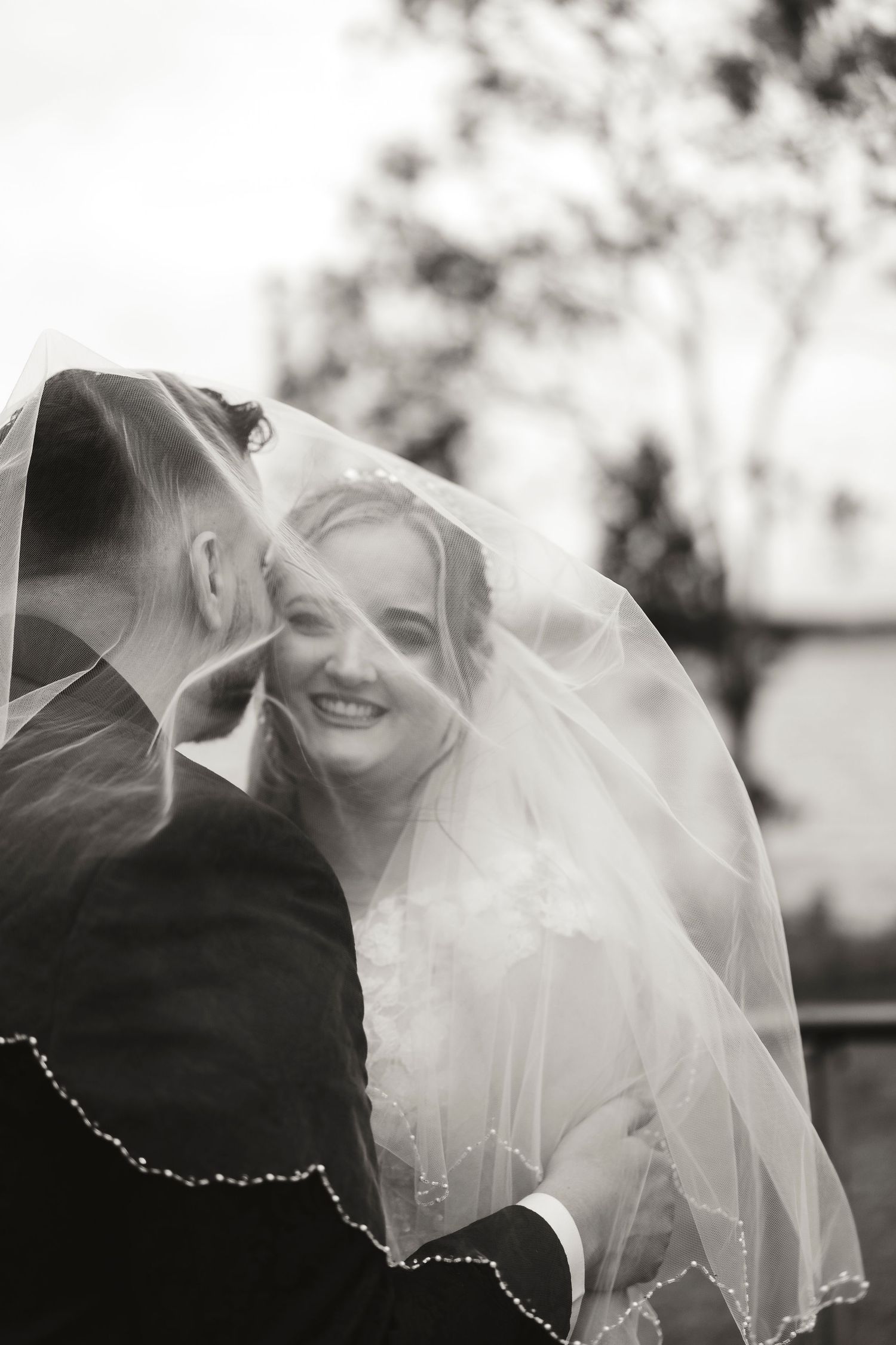 Black and white wedding photo of smiling bride under veil with groom, romantic outdoor portrait.